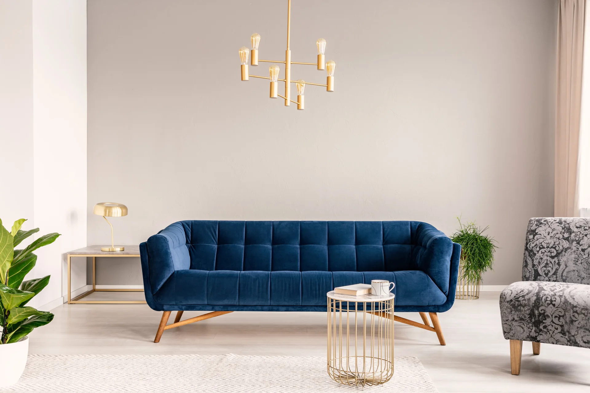 A modern living room features a blue tufted velvet sofa, a patterned gray armchair, a gold wire coffee table, and a brass chandelier.