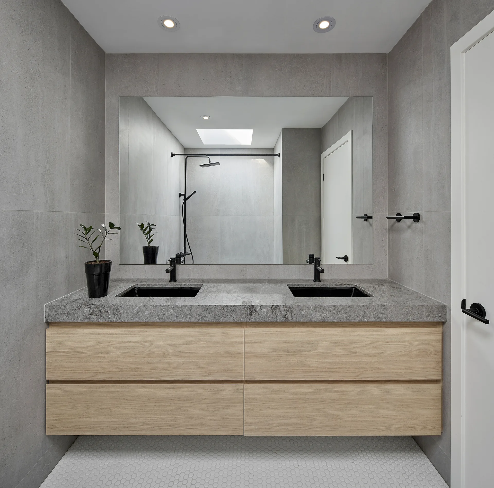 A modern bathroom features a floating light wood double vanity with grey stone countertops, black undermount sinks, and a large frameless mirror.
