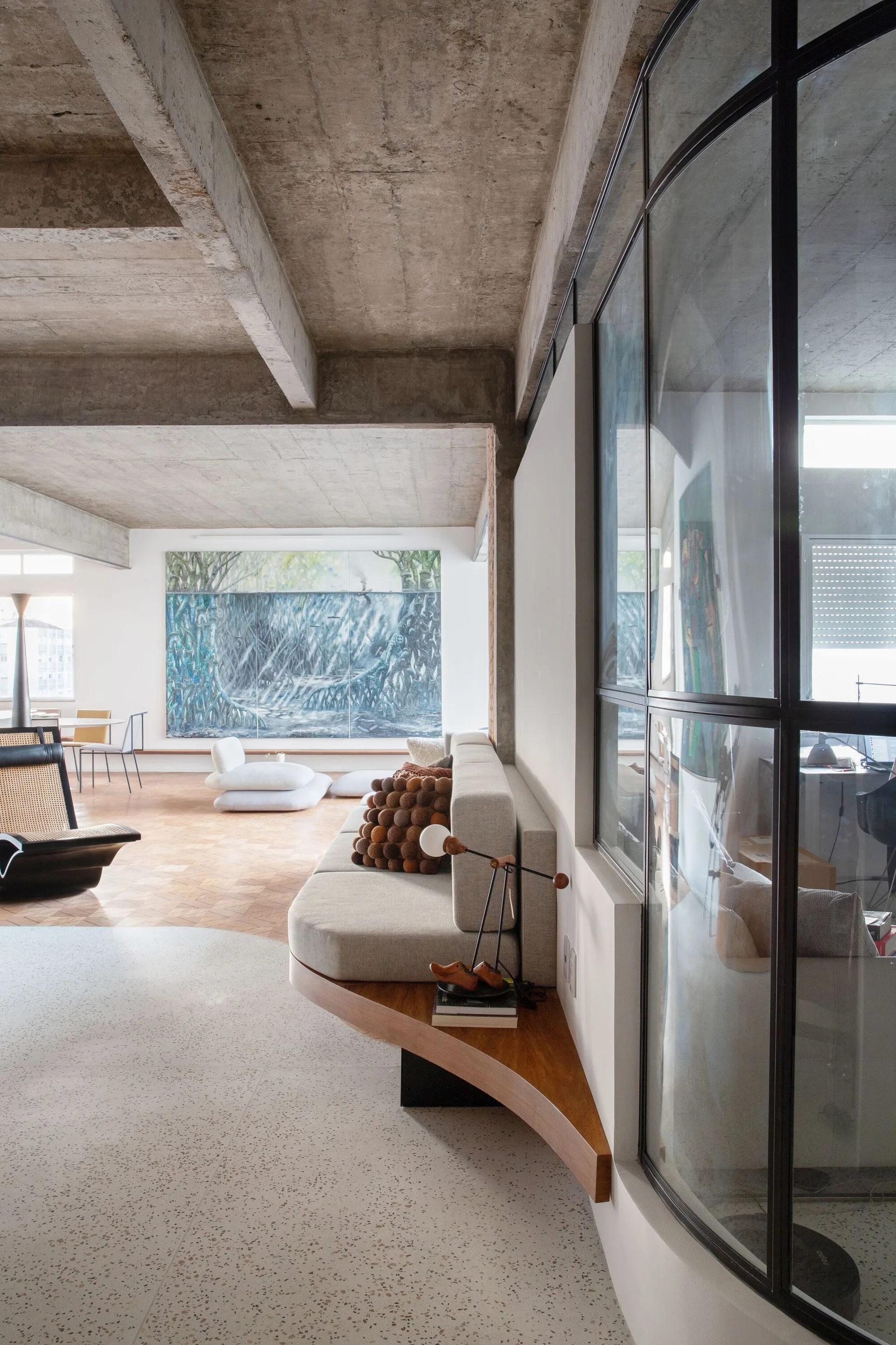 An open-plan living room features a raw concrete ceiling, a curved built-in sofa, and a black metal-framed glass partition wall.