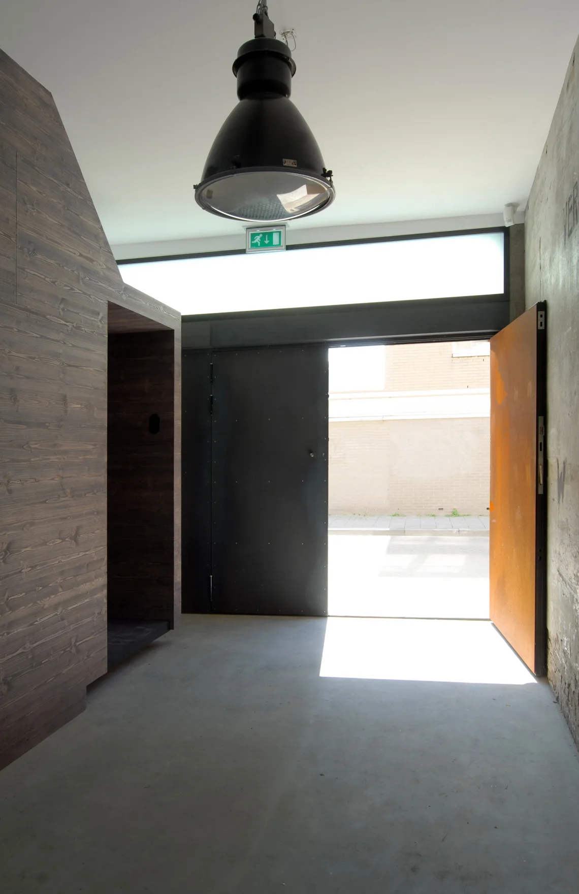 An industrial entry foyer with concrete floors, a dark wood-paneled partition, large metal doors, and a black pendant light.