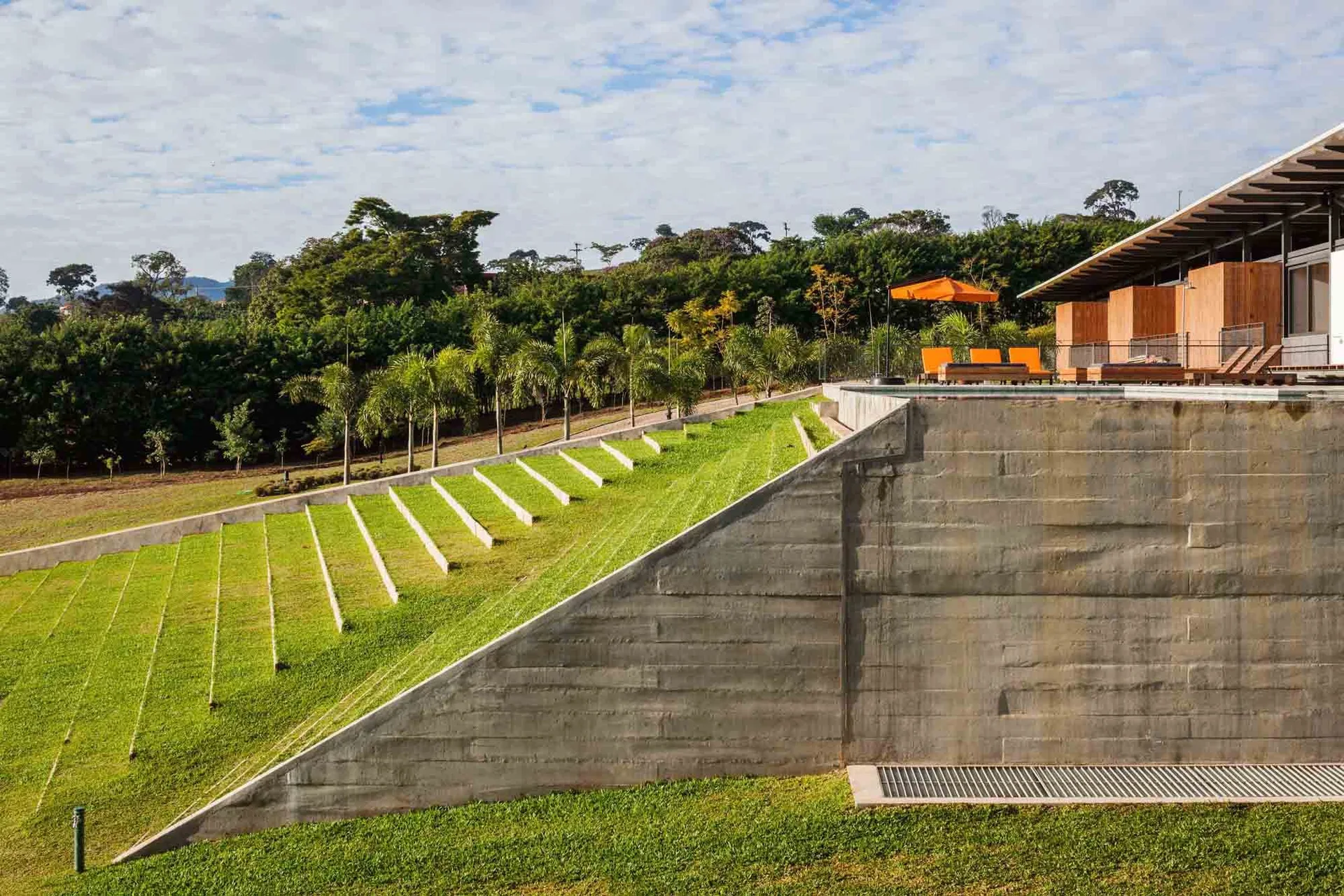 A modern sloped lawn with concrete terrace steps stands next to a tall concrete retaining wall below an elevated pool area with orange lounge chairs.