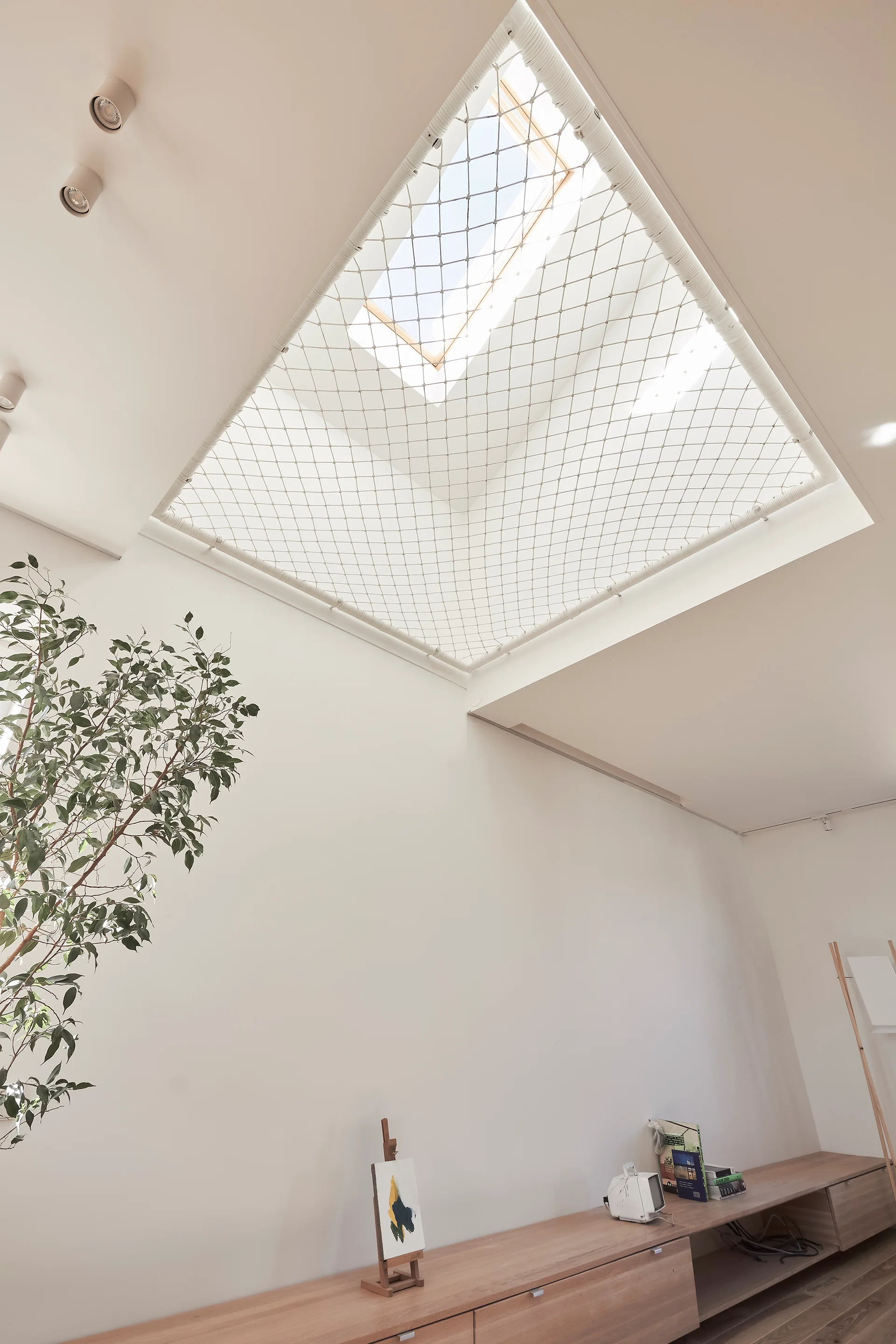 A minimalist off-white room features a large skylight covered with a woven safety net above a low wooden storage console.