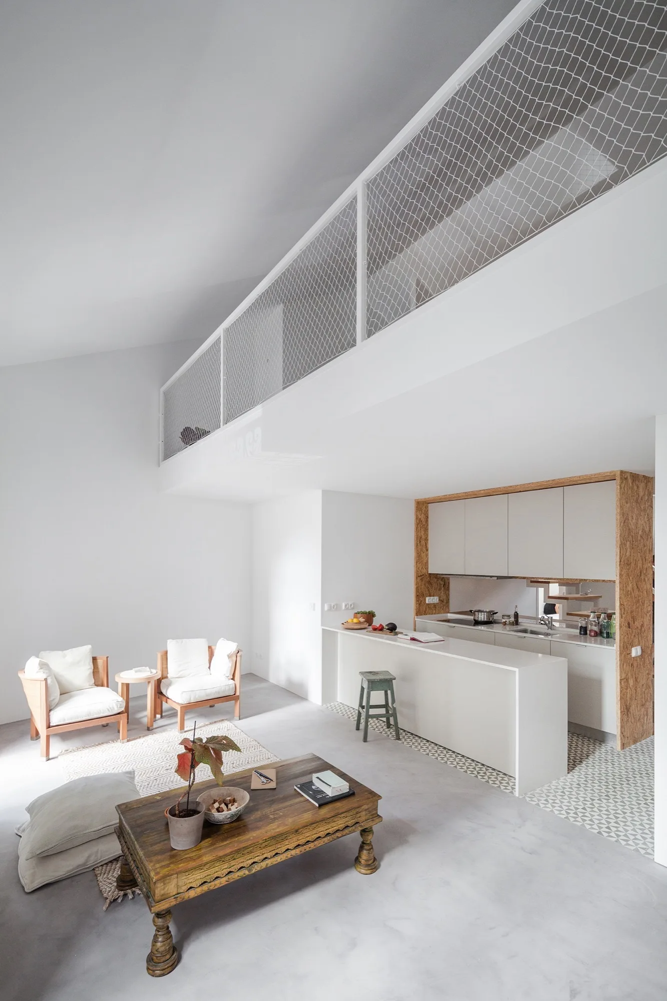 An open-concept living area features wood armchairs, an antique coffee table, and a kitchen with an island beneath a mezzanine loft with white netting.