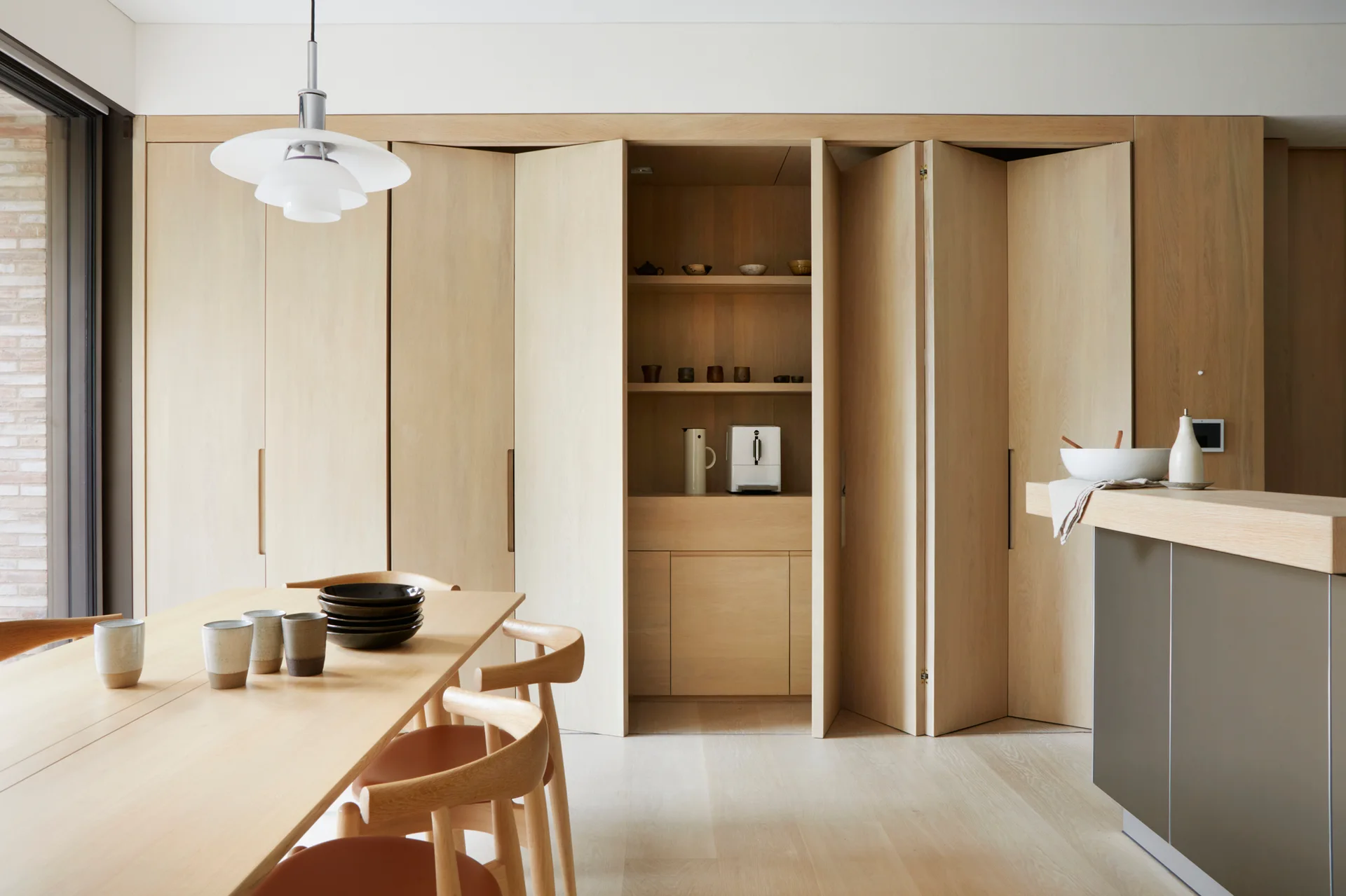A light wood dining table and chairs sit in front of a minimalist kitchen wall featuring full-height bi-fold oak cabinet doors.