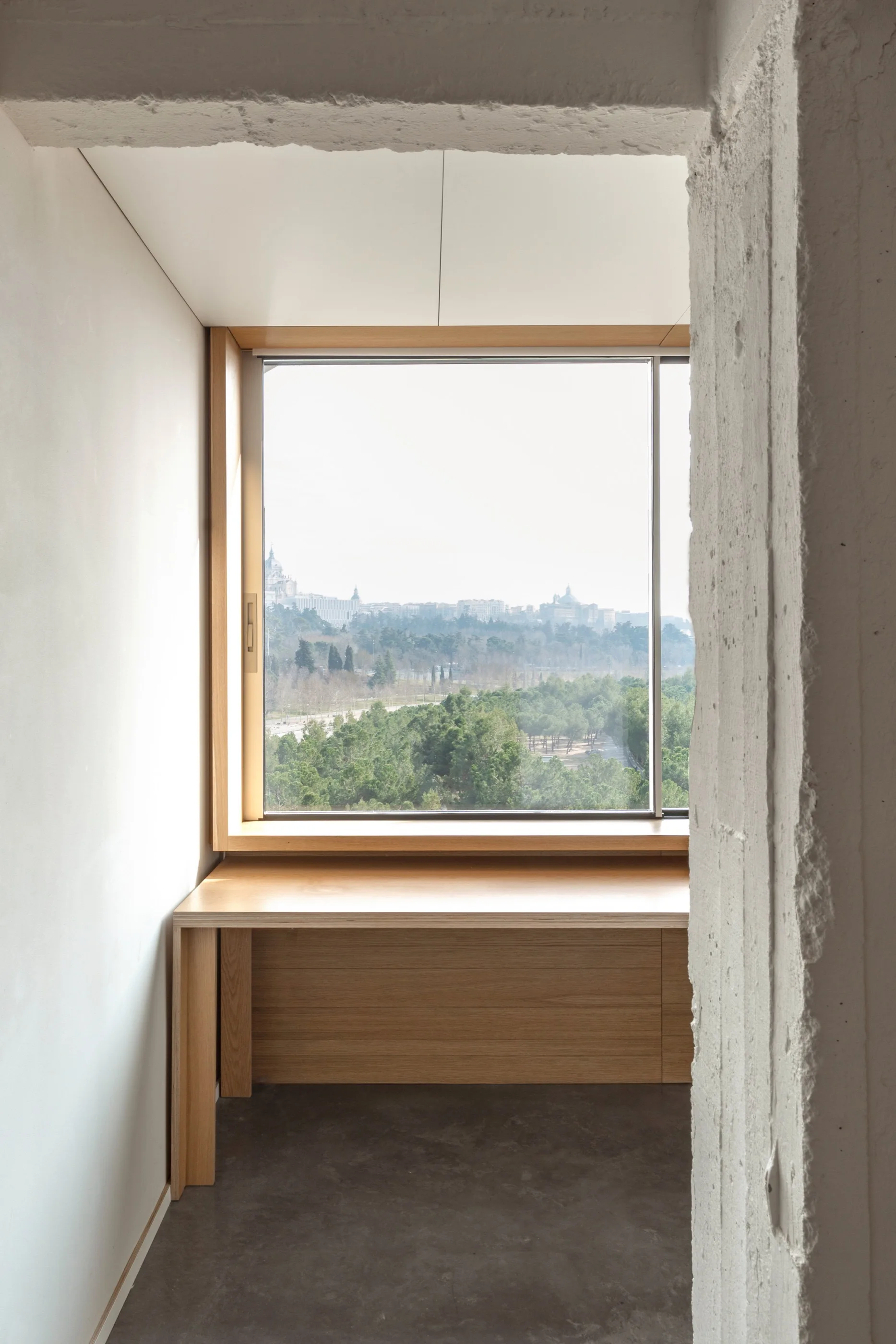 A minimalist built-in wooden desk is positioned beneath a large window framing a tree-filled landscape next to a textured concrete wall.