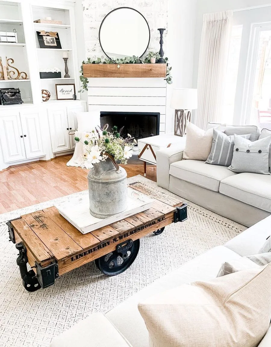 A bright farmhouse living room featuring an industrial cart coffee table, an off-white sofa with accent pillows, and a shiplap fireplace.