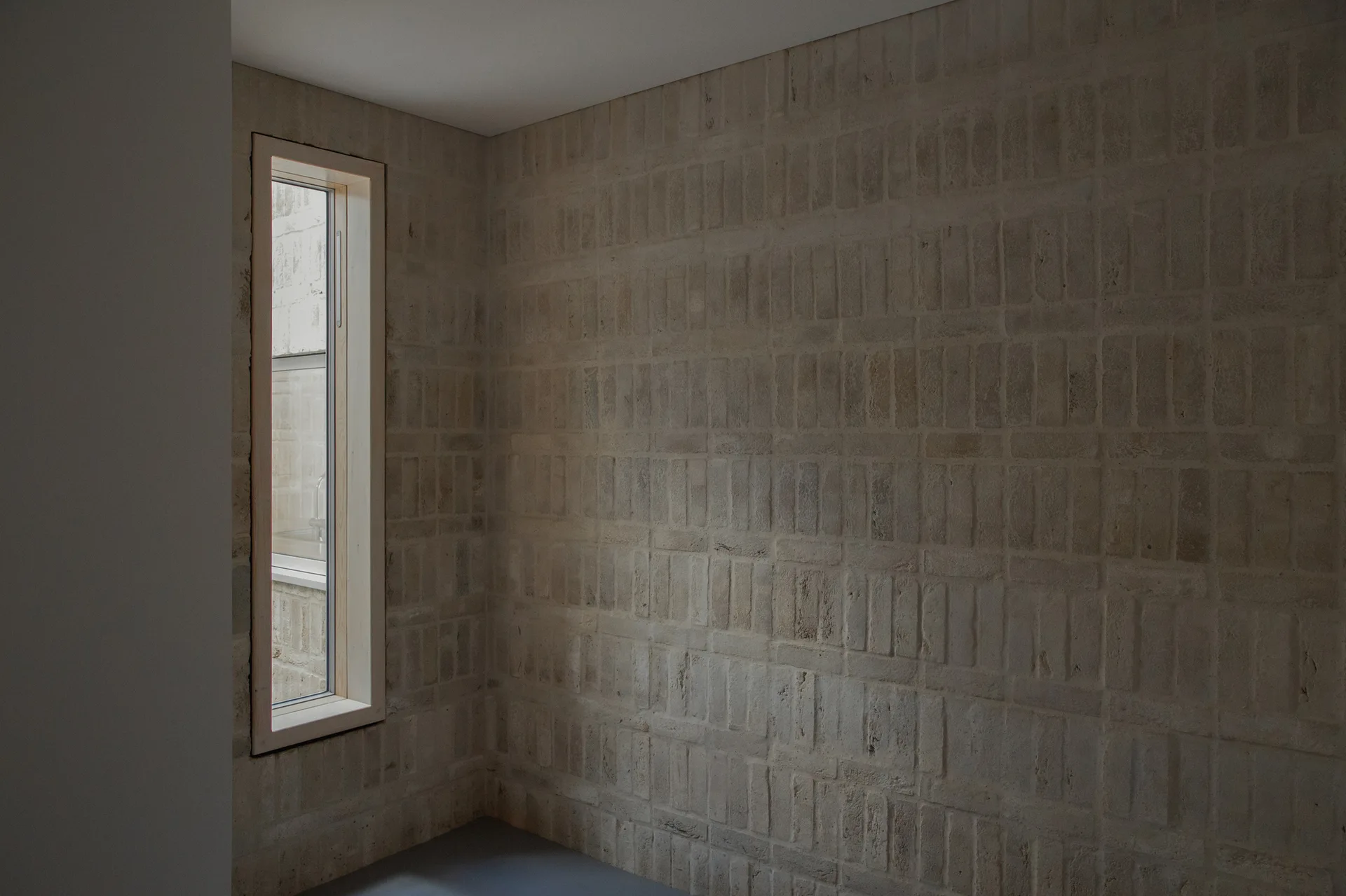 An interior corner features a textured beige brick wall next to a tall, narrow vertical window with a light wood frame.