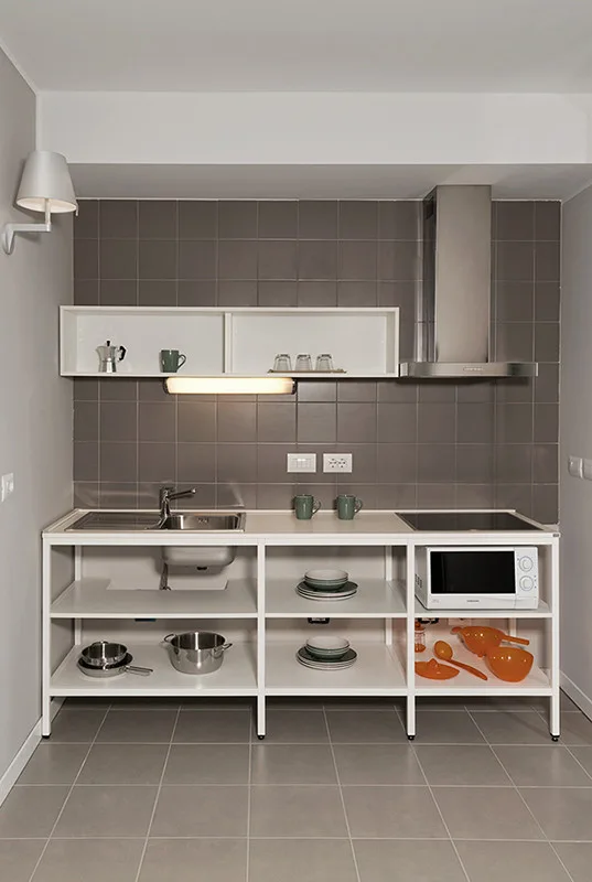 A minimalist kitchen features a white metal open shelving base, a grey square tiled backsplash, and stainless steel appliances.