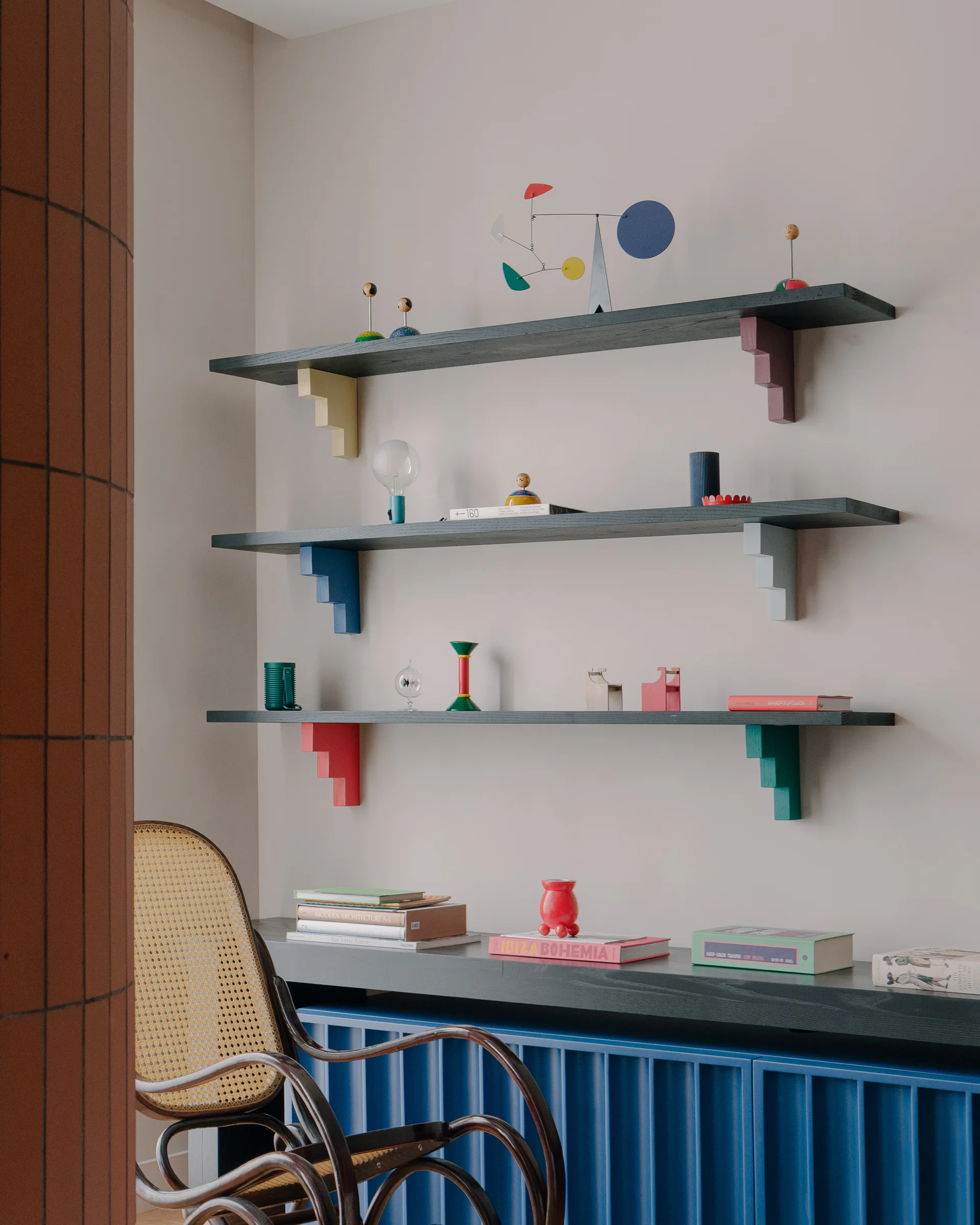 Three black wall shelves with colorful stepped brackets sit above a blue fluted cabinet next to a classic bentwood rocking chair.