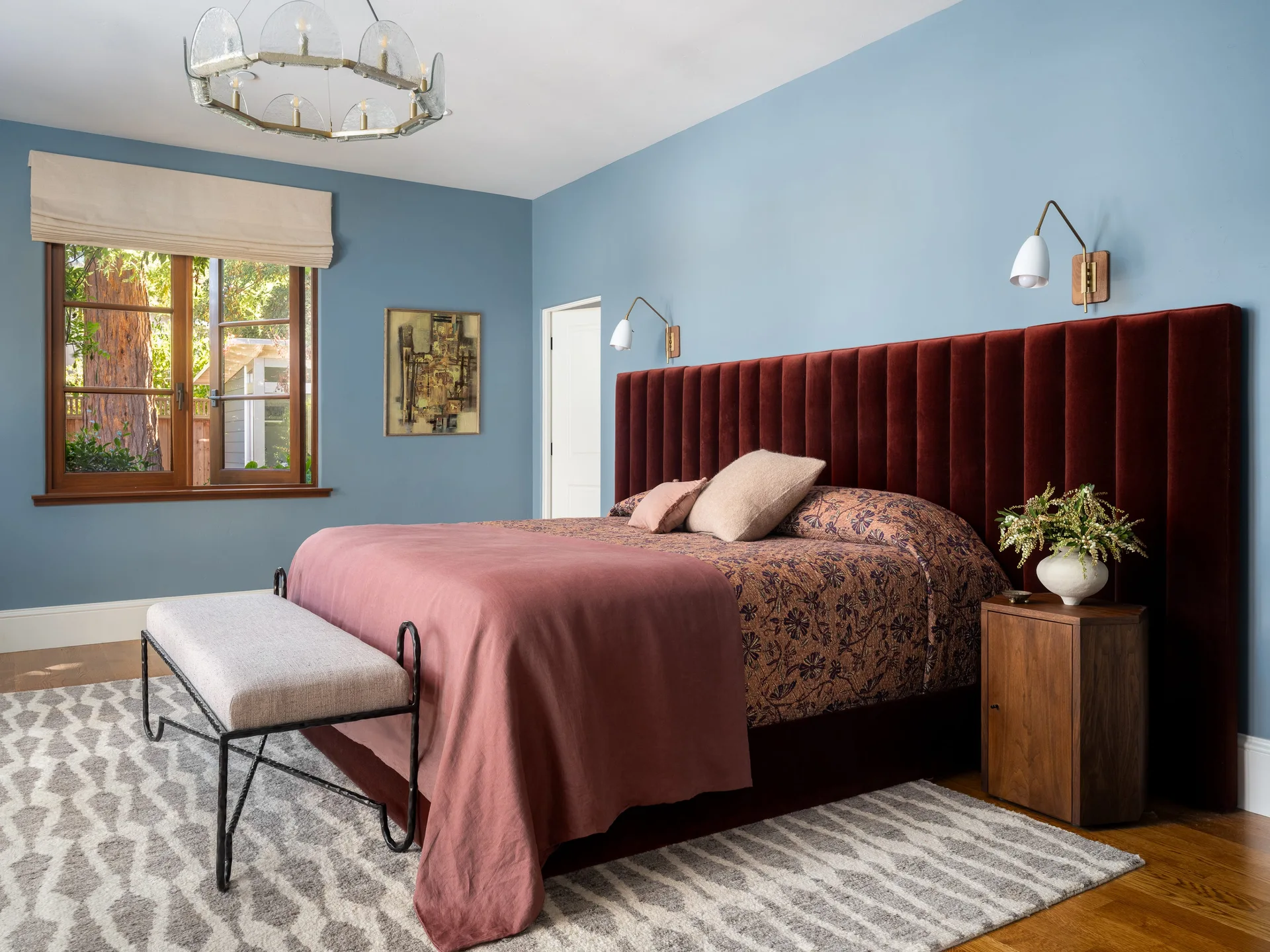 A bedroom featuring a large burgundy velvet channel-tufted headboard against blue walls, a patterned bedcover, a wood nightstand, and a geometric rug.