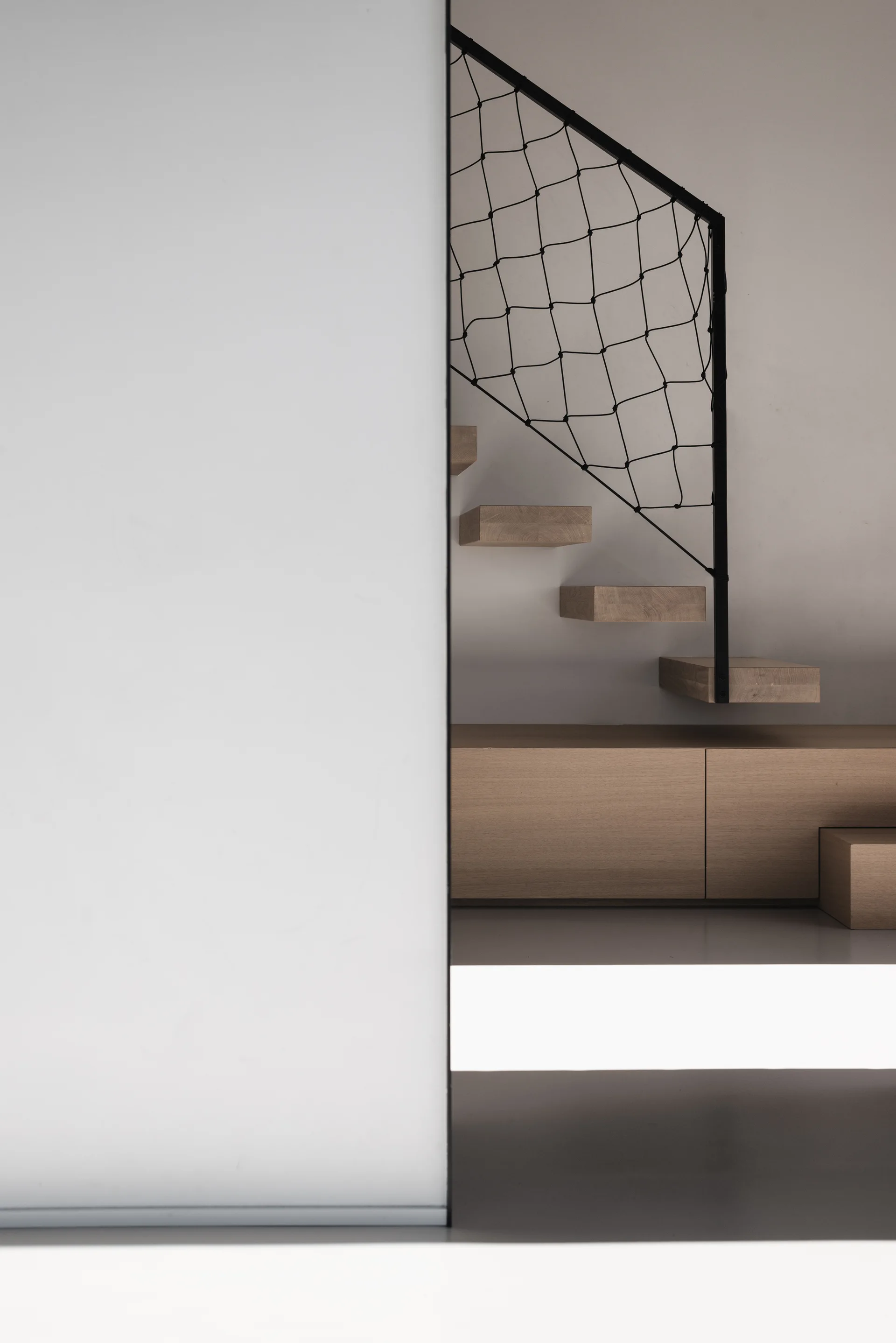 A minimalist floating wood staircase features black metal mesh railing above custom light oak storage cabinetry.