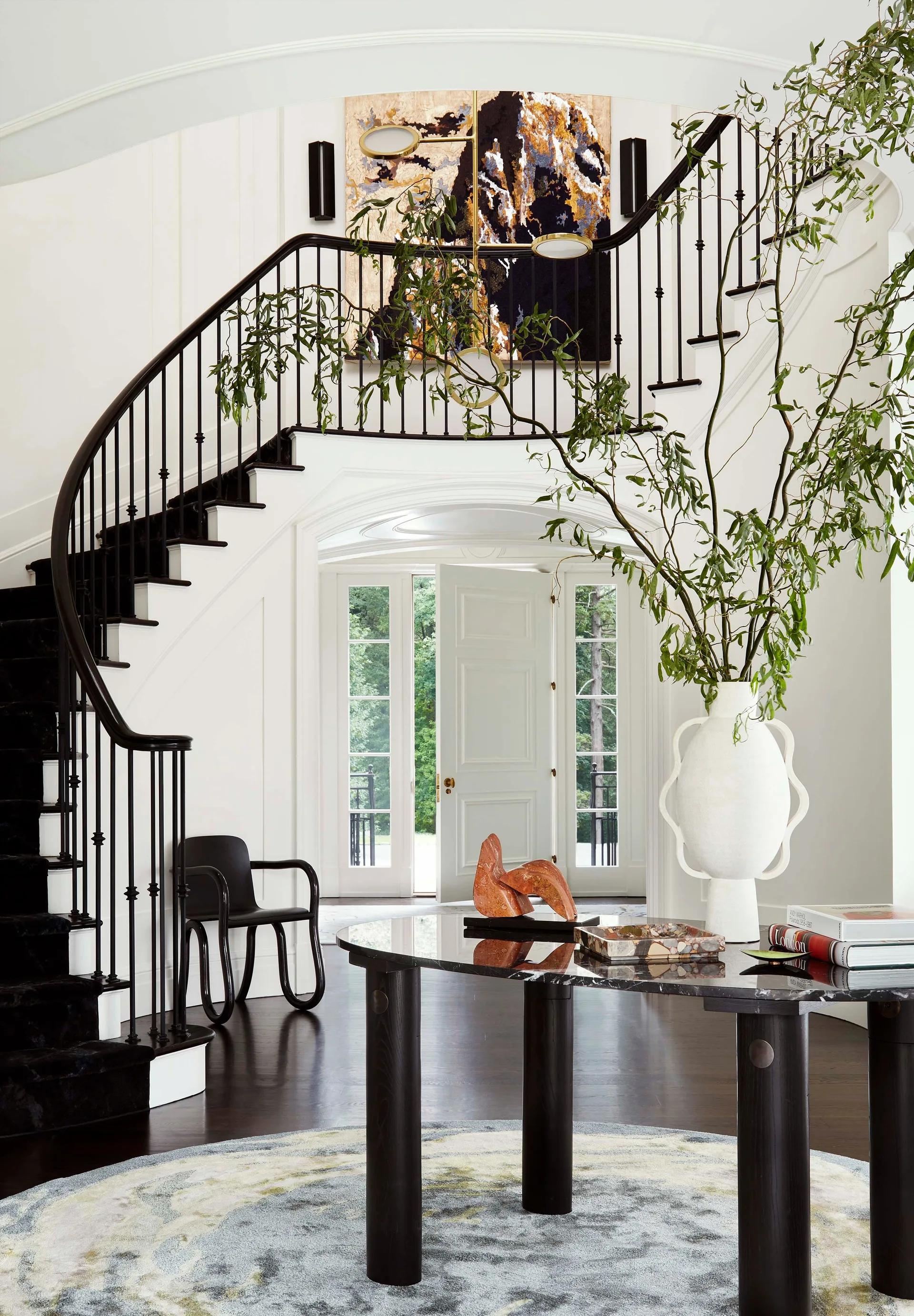 A bright foyer featuring a curved black staircase with wainscoting, a dark marble-topped console table, and a large white vase with green branches.