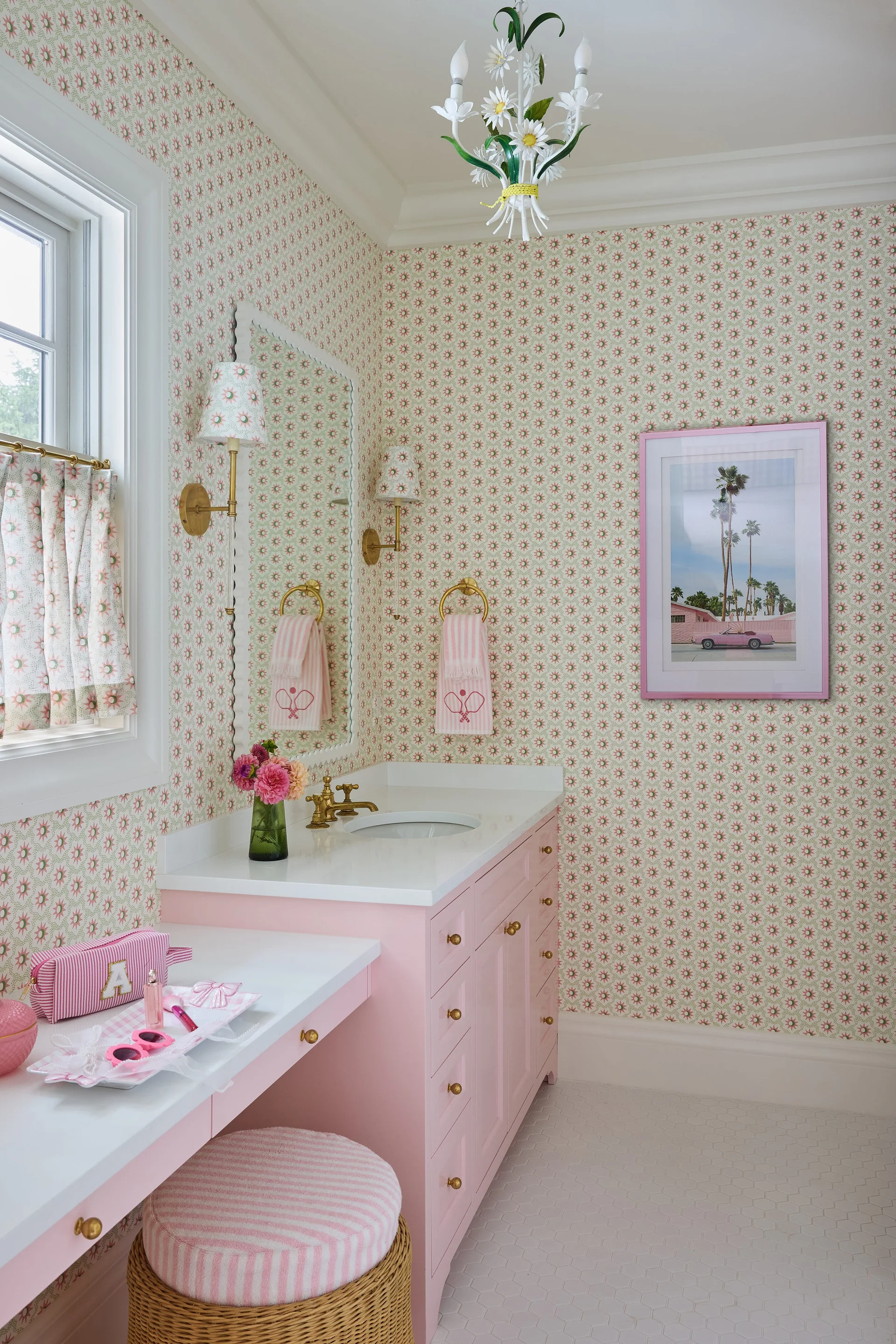 A bathroom featuring a pastel pink vanity under a scalloped mirror, floral wallpaper, and a wicker stool with a striped cushion.