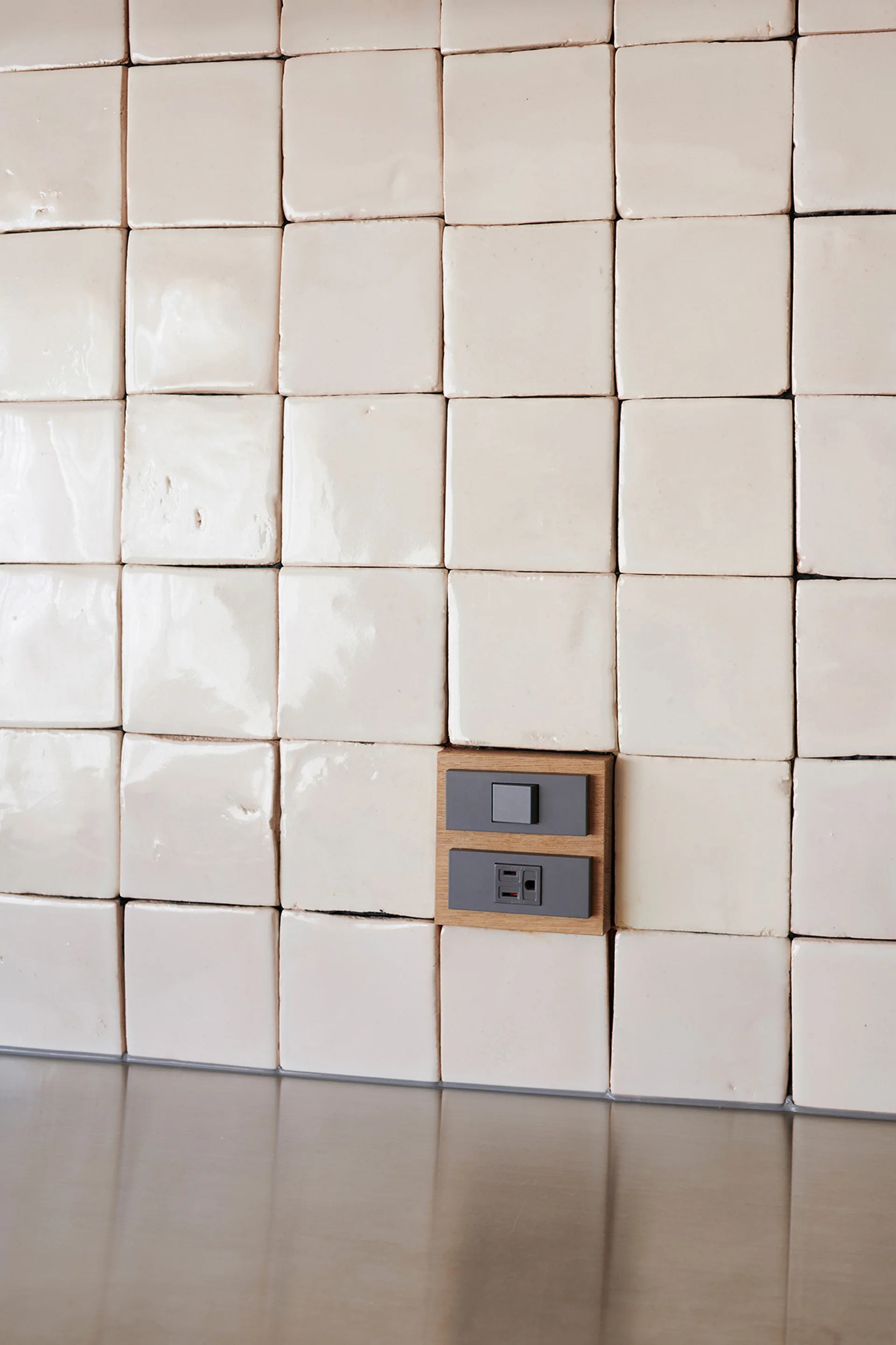 A close-up of glossy off-white square ceramic wall tiles arranged in a grid above a metal countertop, featuring an integrated grey outlet in a wood frame.