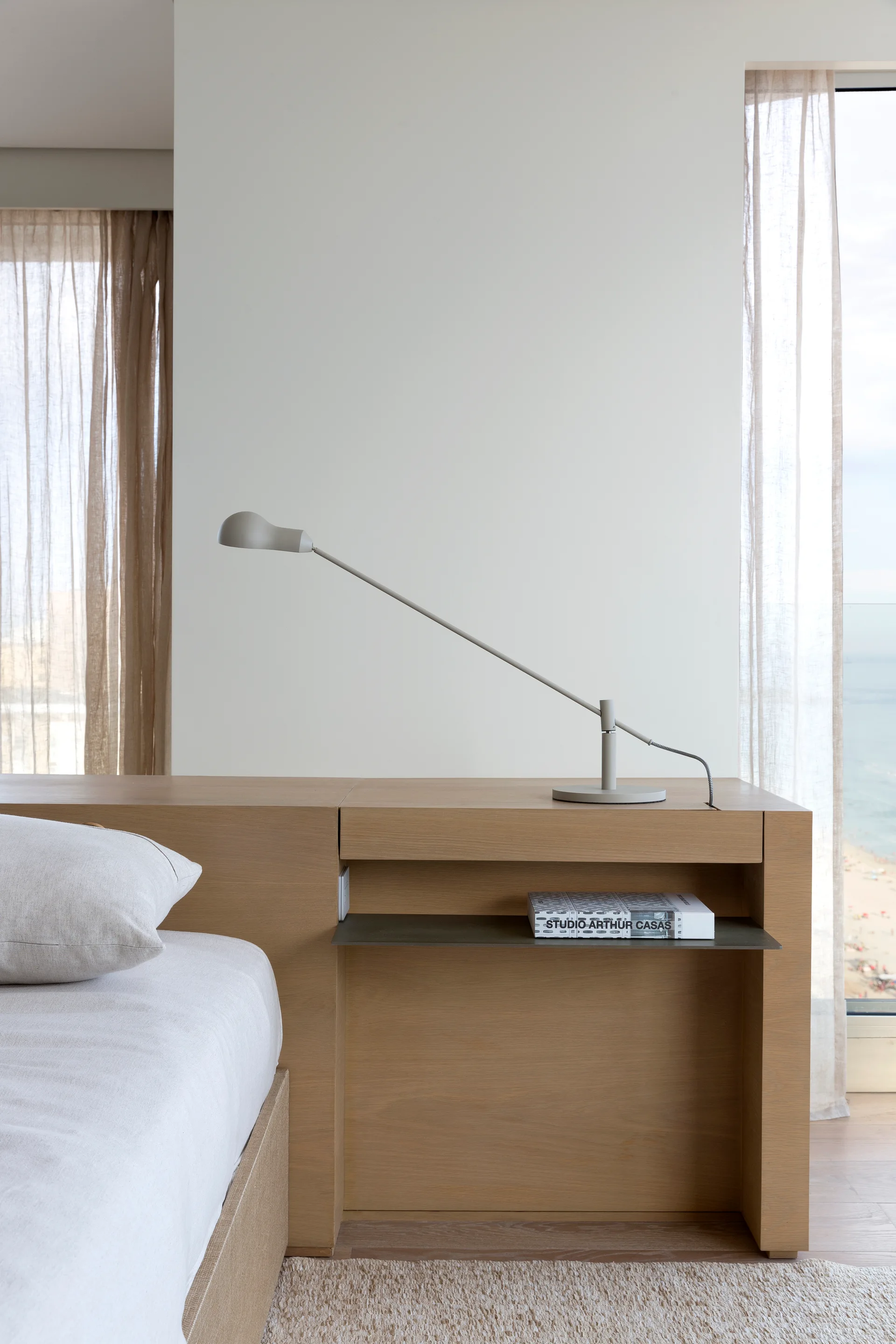 A minimalist bedroom bedside area features a built-in wooden nightstand with a slim metal shelf, a grey task lamp, and sheer curtains.