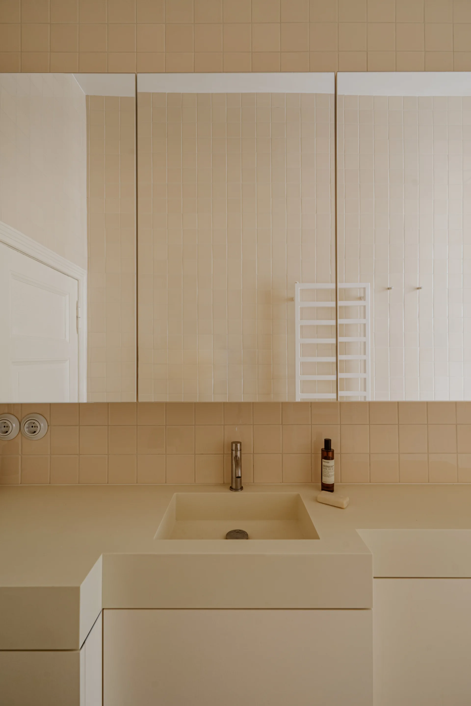 A monochromatic beige bathroom vanity with an integrated sink, a large mirror, and walls covered in small square tiles.