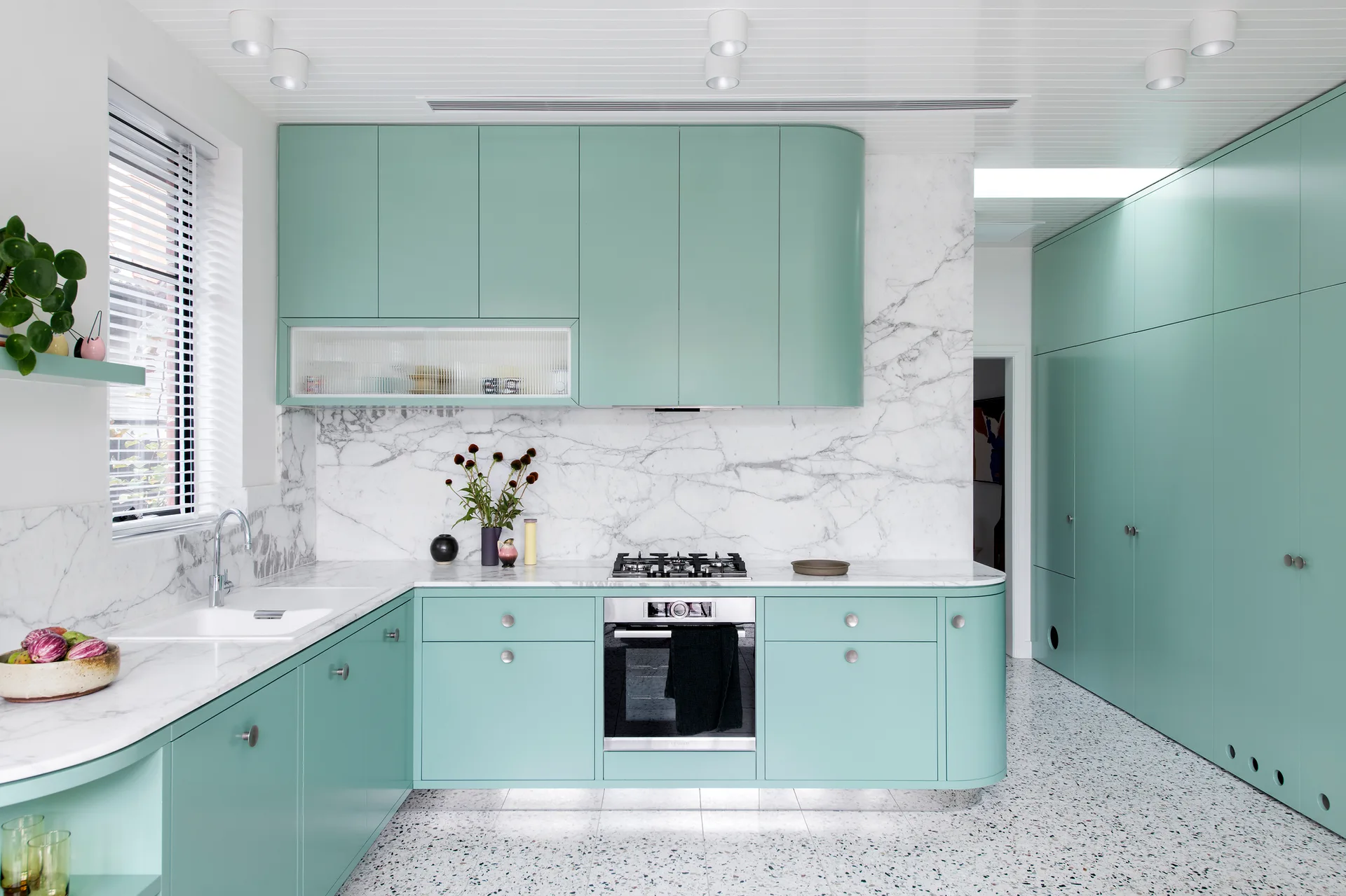 A modern kitchen features curved mint green cabinetry, a white marble countertop and backsplash, and speckled terrazzo tile flooring.