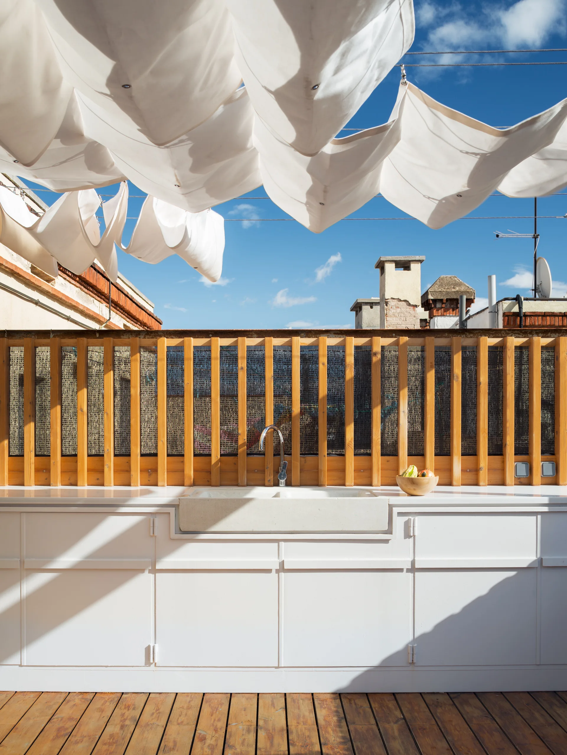 An outdoor kitchen on a wood deck features white cabinets, a stone sink, a vertical wood slat privacy screen, and white canopy fabric overhead.