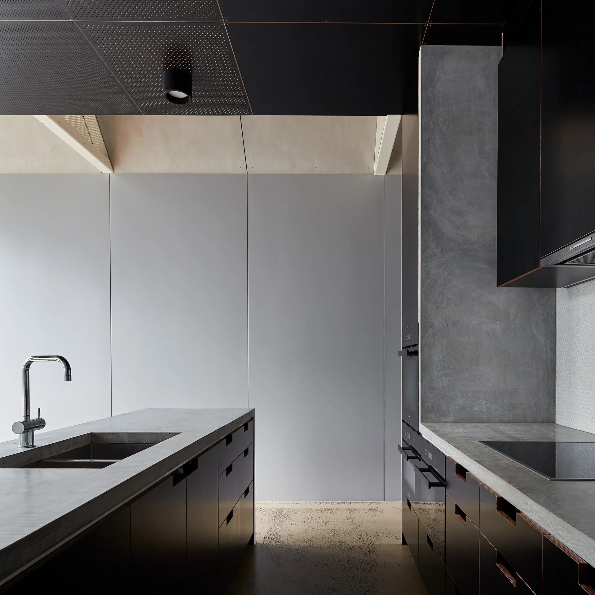 A modern kitchen features a concrete island with a stainless steel sink, dark grey cabinetry, integrated appliances, and a perforated black ceiling.