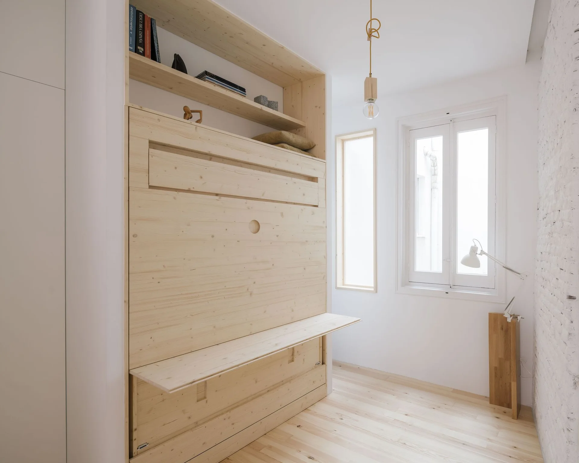 A minimalist white bedroom features an integrated light wood Murphy bed wall unit with built-in shelves, a matching desk, and wood flooring.