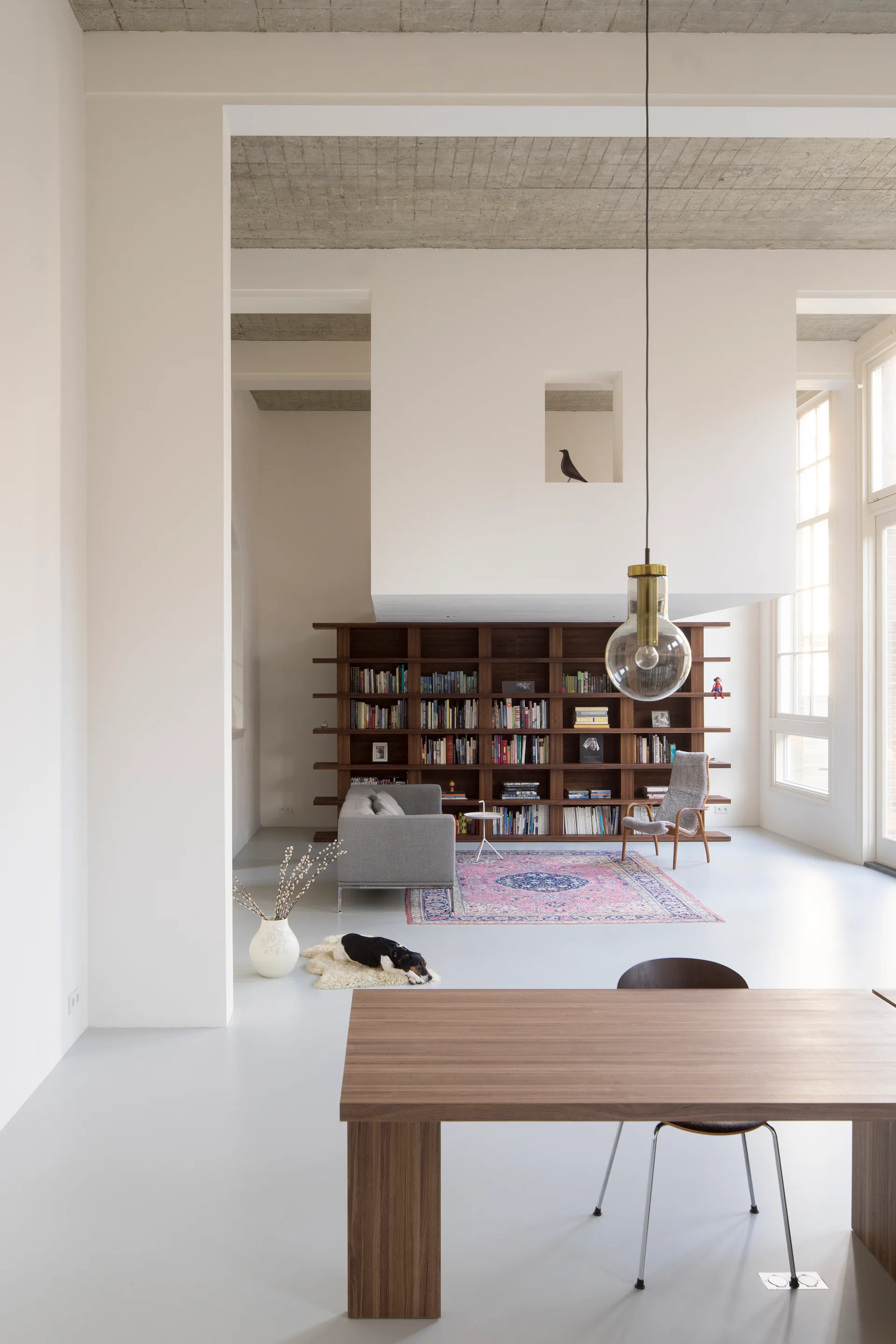 A minimalist open living room featuring an exposed concrete ceiling, a large dark wood bookshelf against a white wall, and a pink patterned rug.