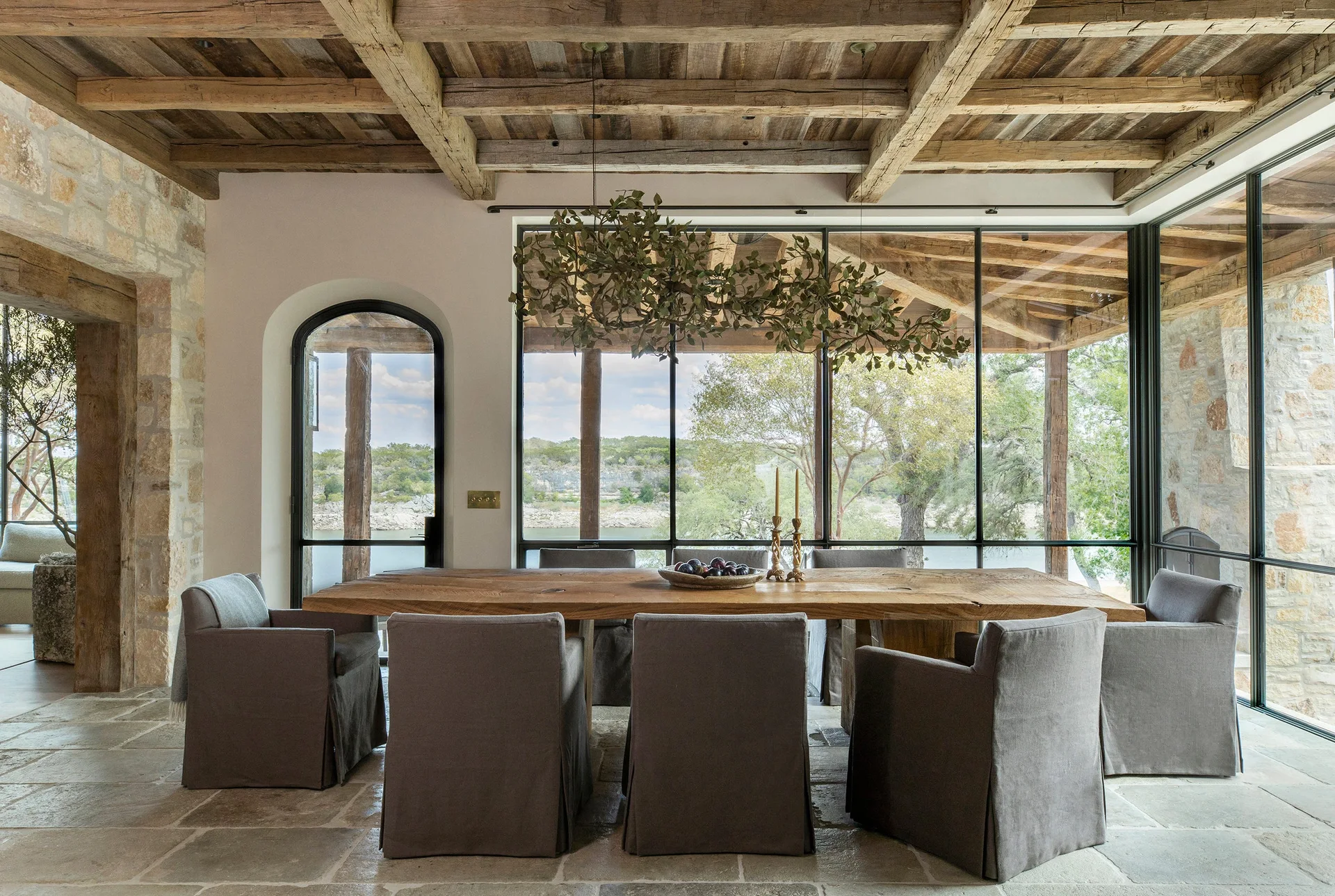 A rustic dining room features a long wooden table, grey slipcovered armchairs, an exposed beam ceiling, and large windows.
