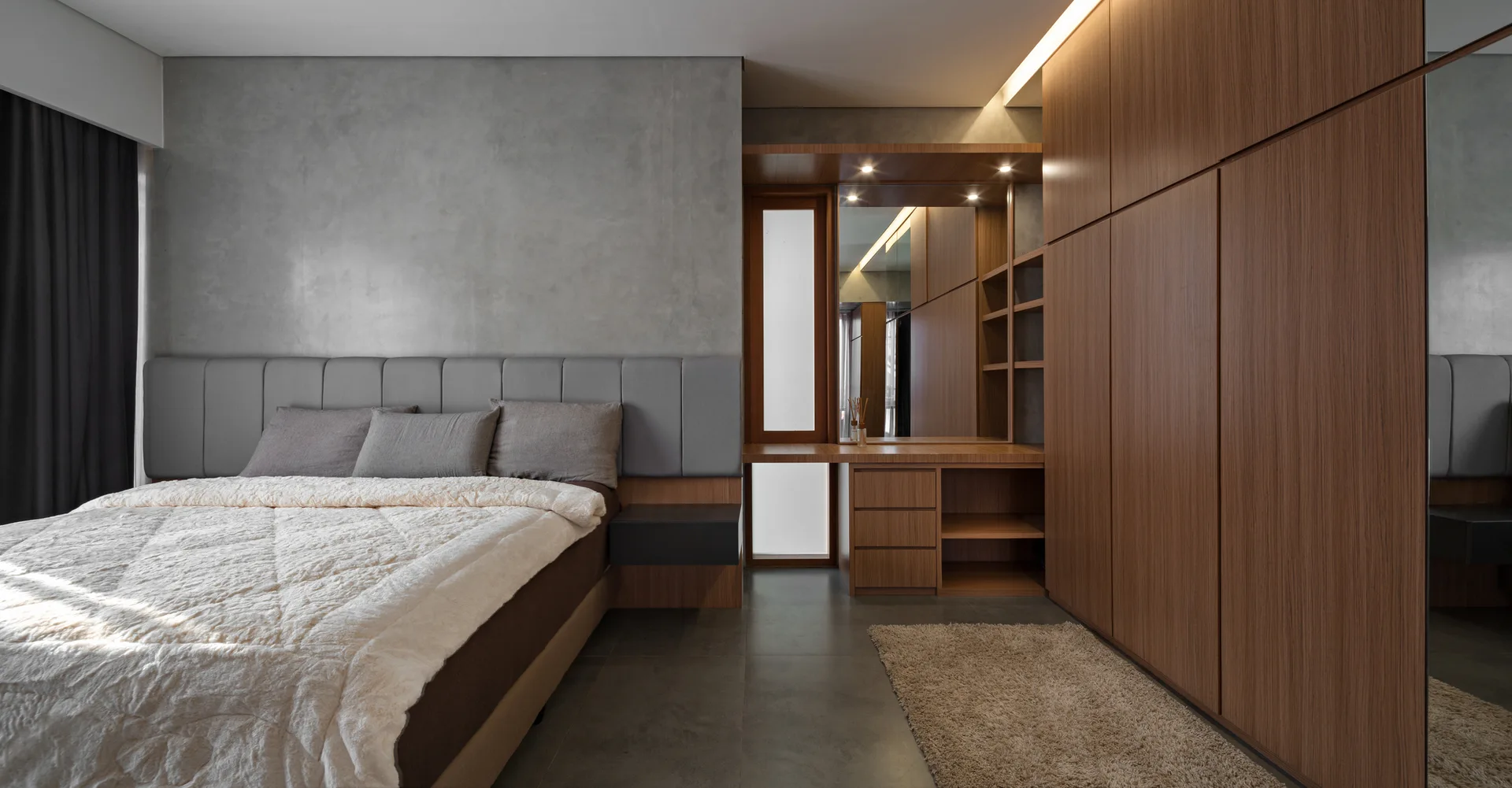 A modern bedroom features an upholstered bed against a grey concrete-textured wall next to a large built-in wood wardrobe and vanity.