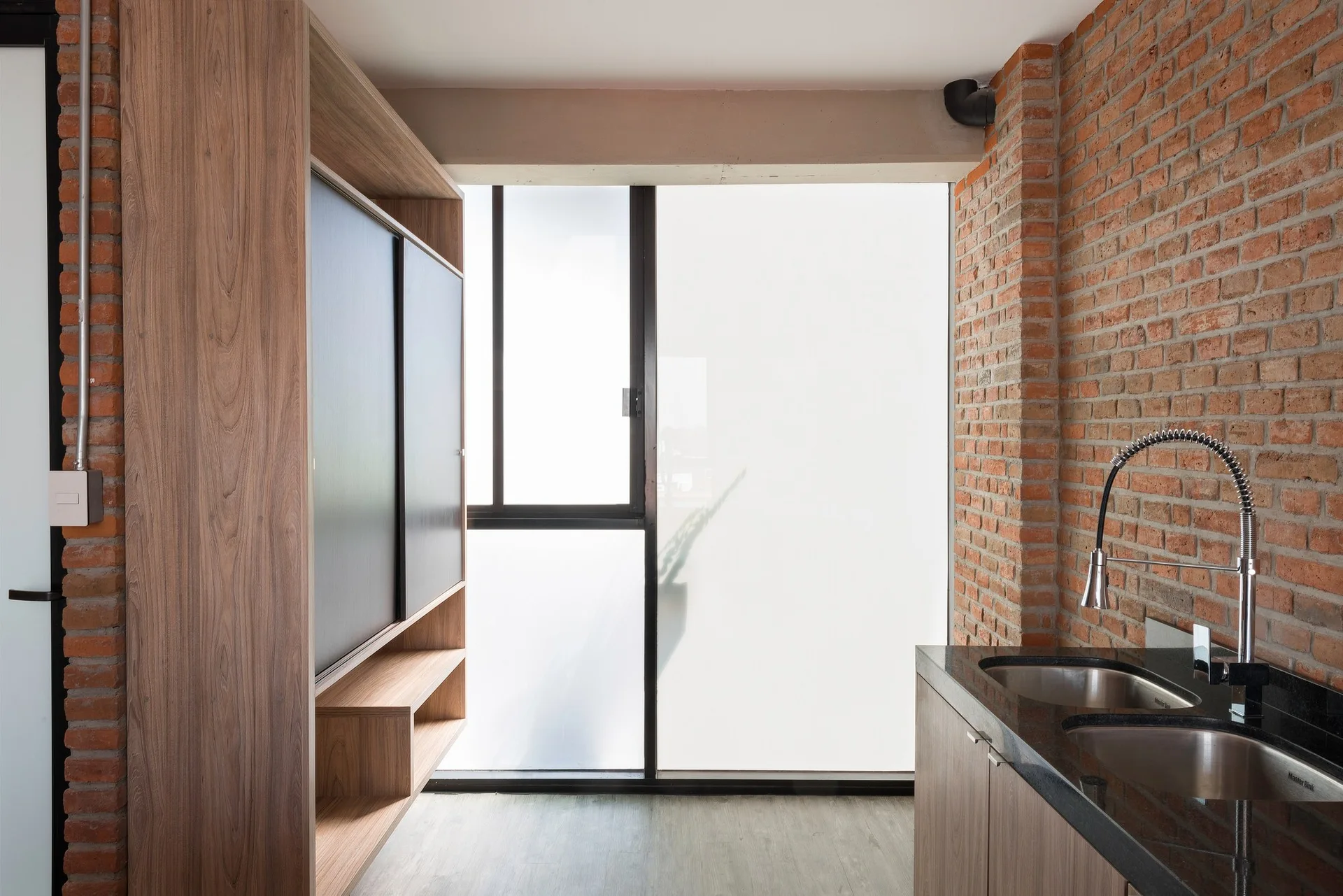 An industrial kitchen features exposed brick walls, a large wooden storage unit, and a black countertop with a double sink under a frosted window.