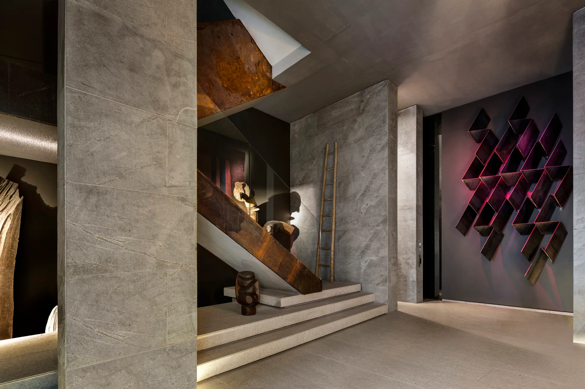 A contemporary gray stone entryway featuring a floating staircase with a rusted metal railing and a geometric wall sculpture backlit with red neon.