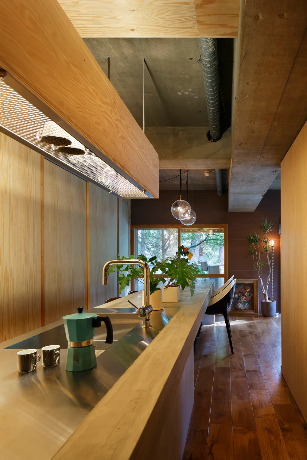 A long wood kitchen island with a stainless steel sink sits beneath a custom wooden light fixture and an exposed concrete ceiling.