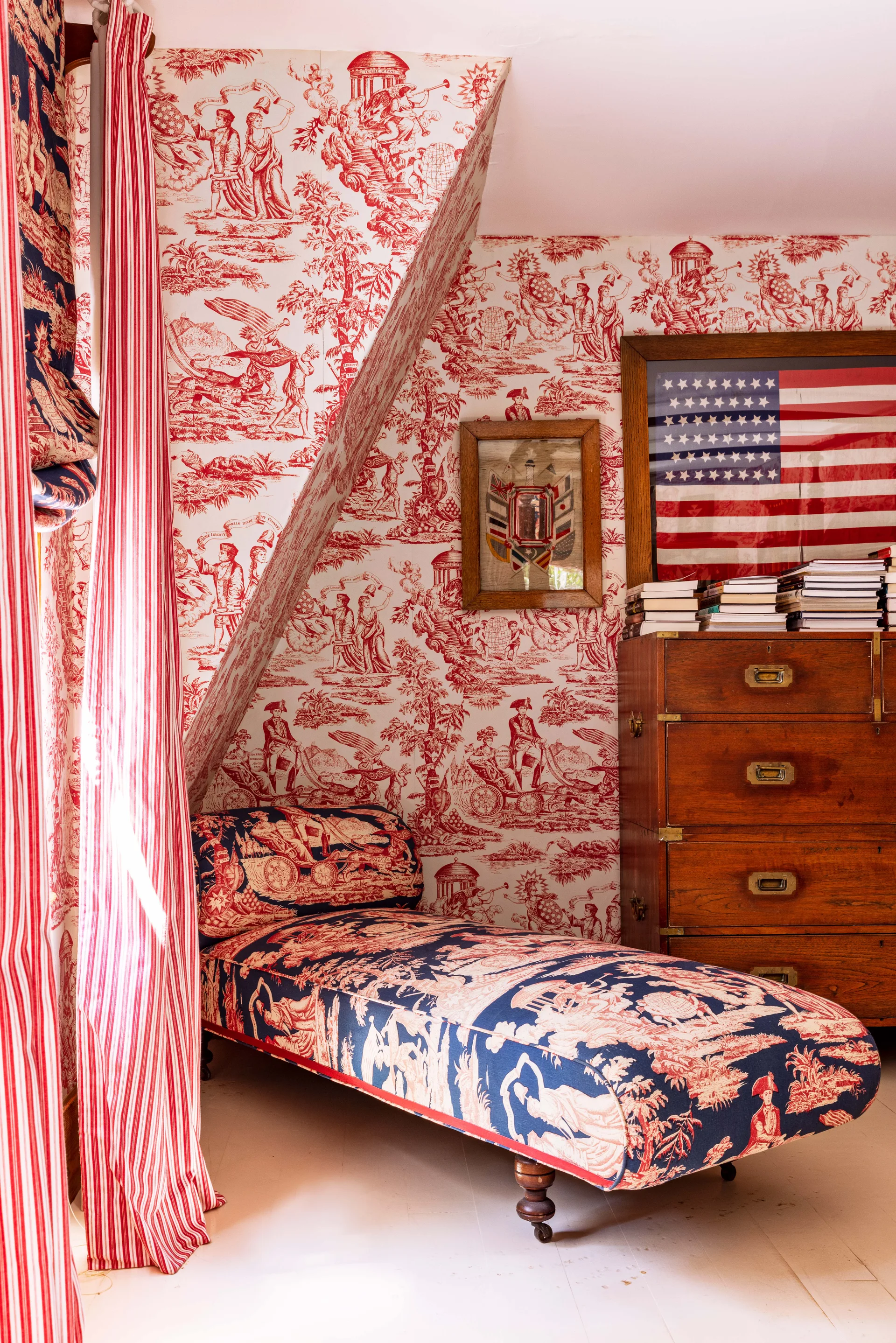 A cozy bedroom corner with red and white toile wallpaper, a blue and red patterned chaise lounge, and a wooden chest of drawers.