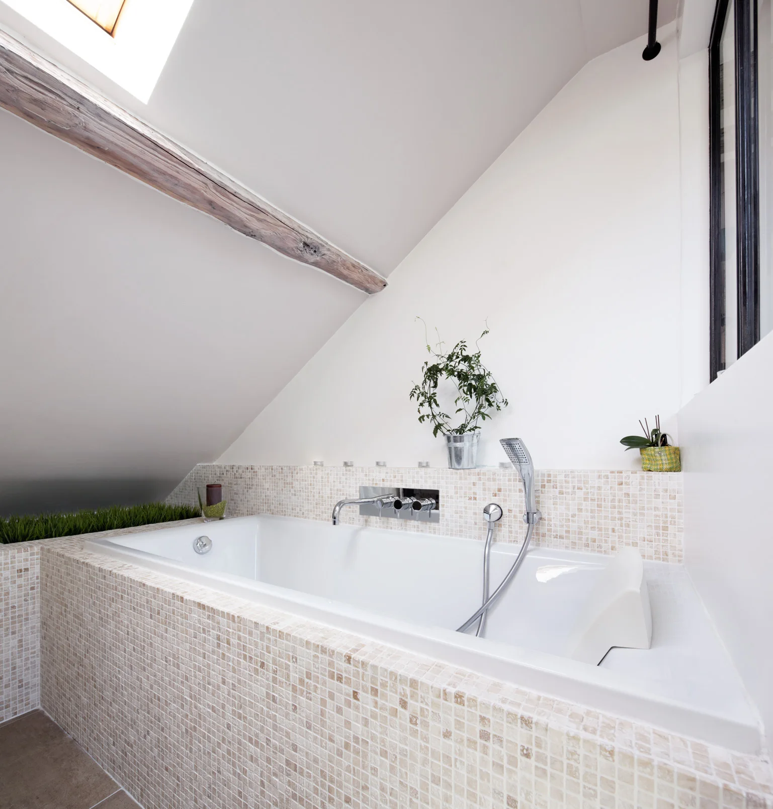 A built-in white bathtub with a beige mosaic tile surround sits beneath a slanted attic ceiling with an exposed wooden beam.