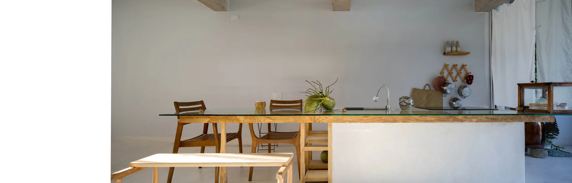 A minimalist kitchen features a white island with a light wood frame, a glass countertop extending into a dining table, and wooden chairs.
