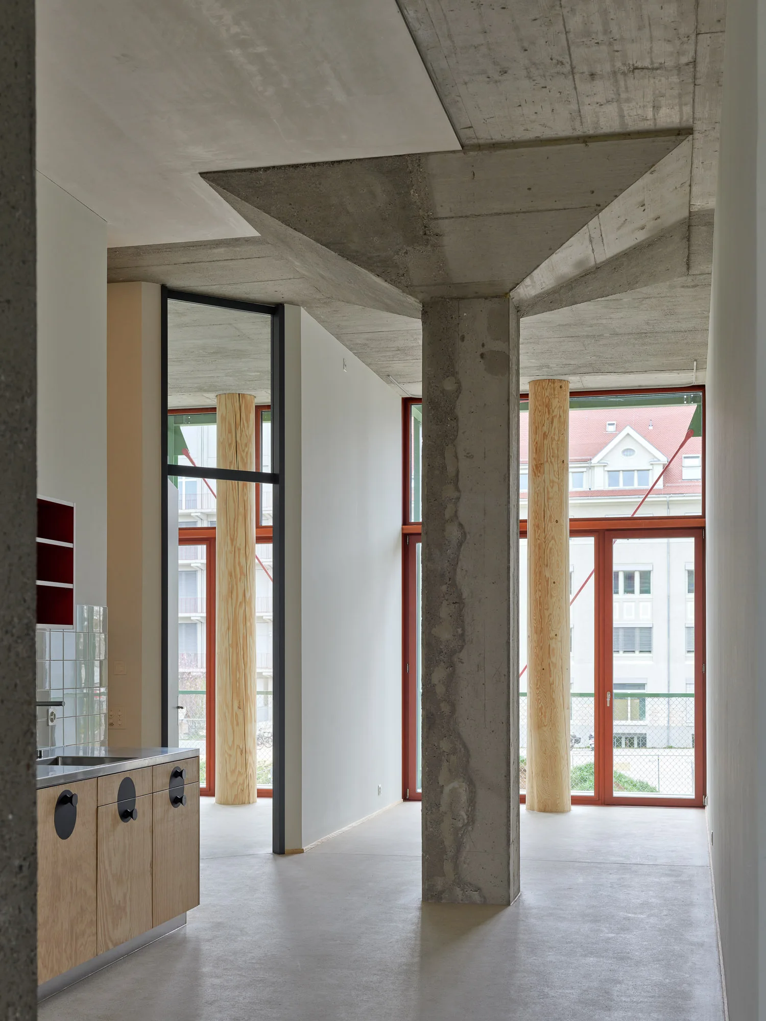A modern industrial loft space features a raw concrete structural column, light wood kitchen cabinets, and cylindrical wood pillars in front of large windows.
