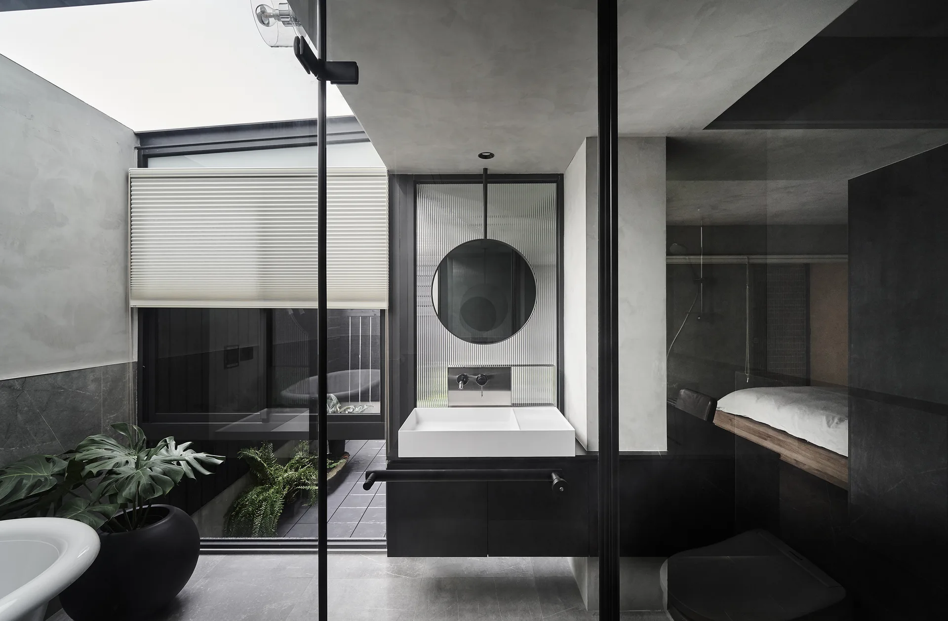 A modern grey bathroom with a black floating vanity, circular mirror, fluted glass panel, and large glass walls overlooking a courtyard.