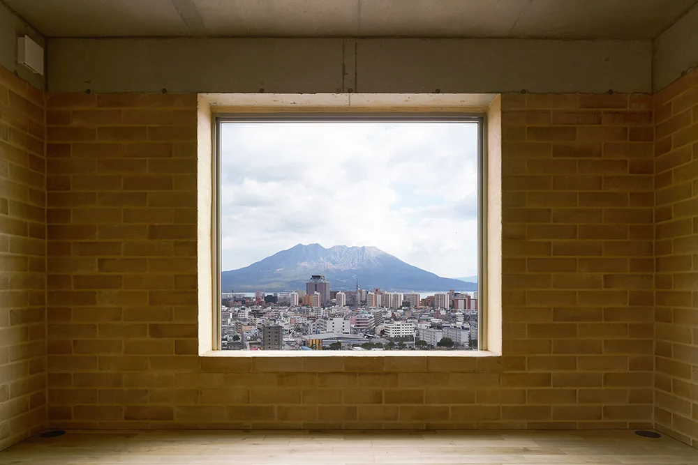 A minimalist interior features warm tan brick walls, a raw concrete ceiling, and a large square window framing a city and mountain landscape.