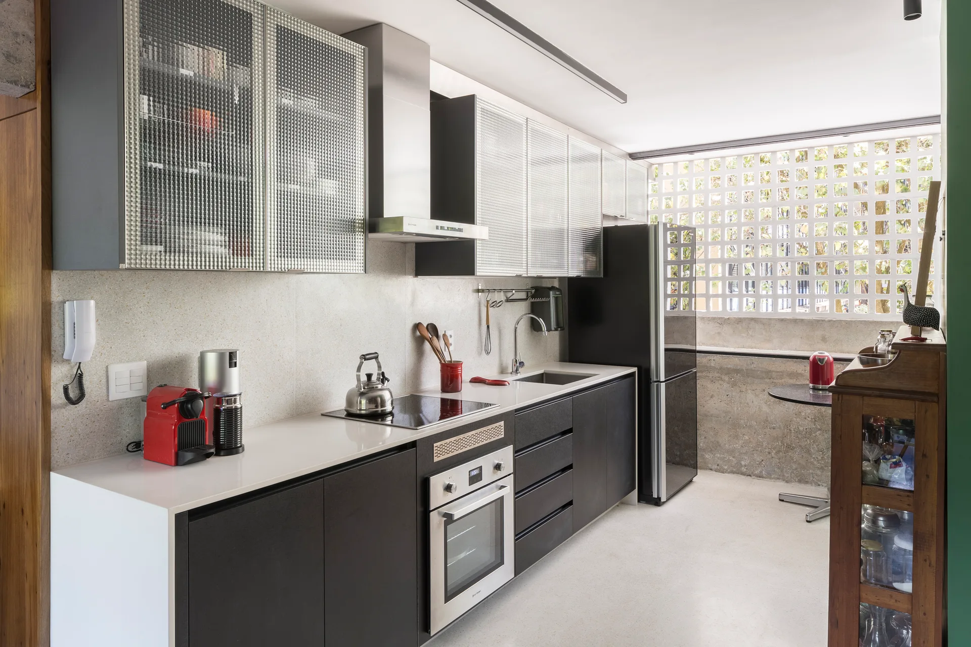 A modern kitchen featuring black lower cabinets, upper cabinets with textured glass fronts, and a geometric concrete lattice window at the far end.