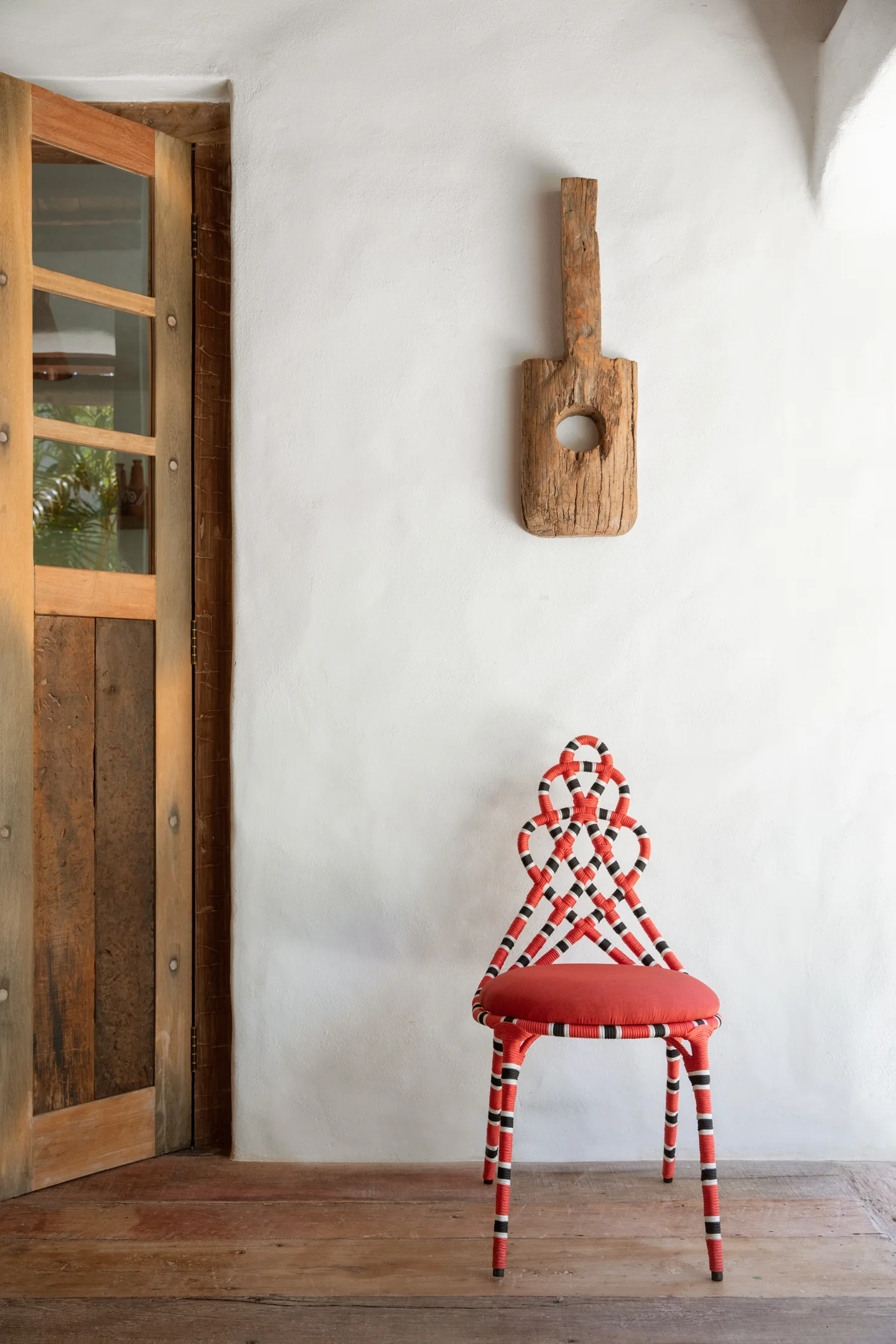 A red and black woven sculptural chair stands on a wooden floor against a white plaster wall next to an open wooden door.