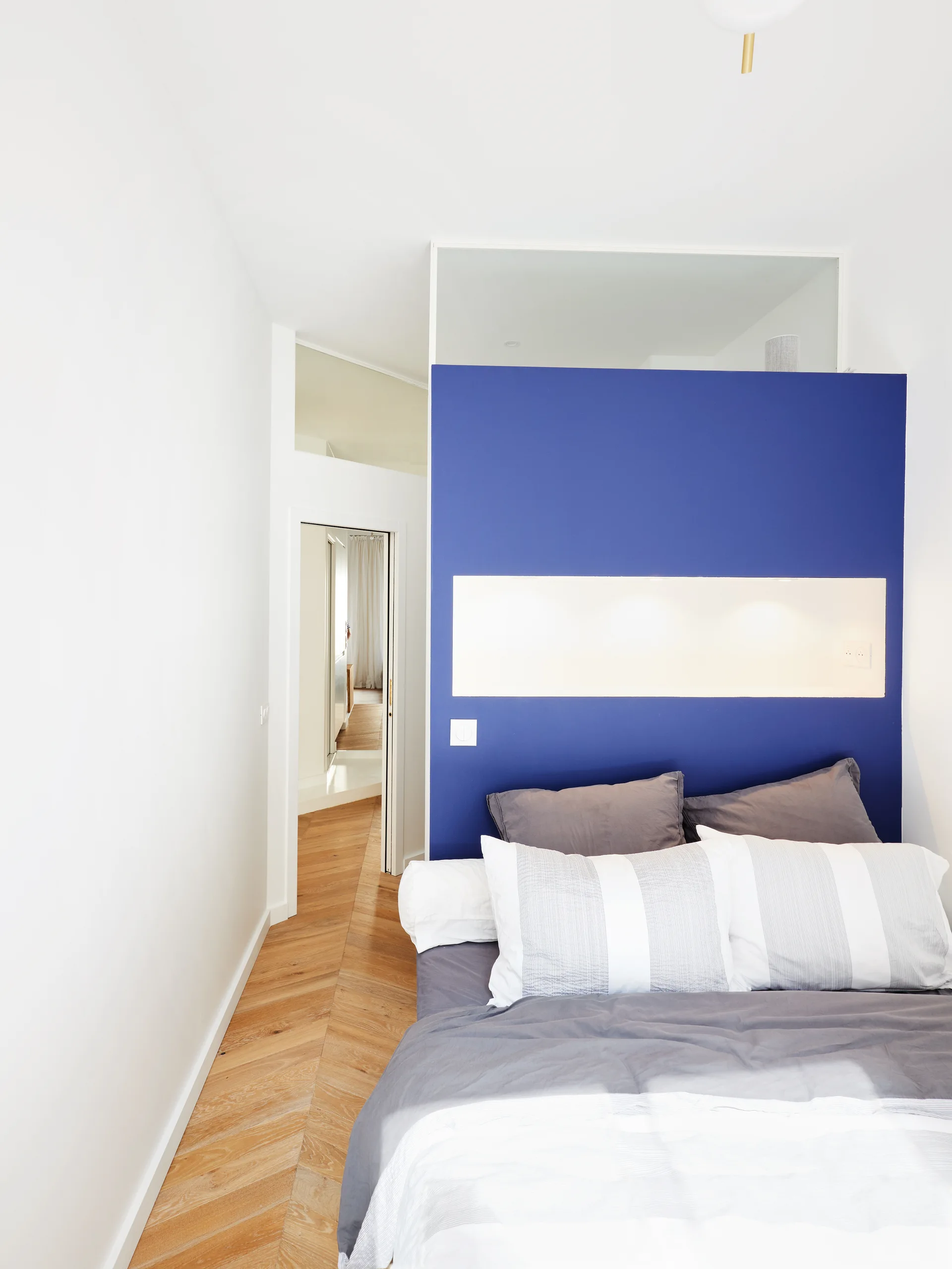 A modern bedroom features a bright blue partition wall behind a bed with a built-in illuminated niche and herringbone wood flooring.
