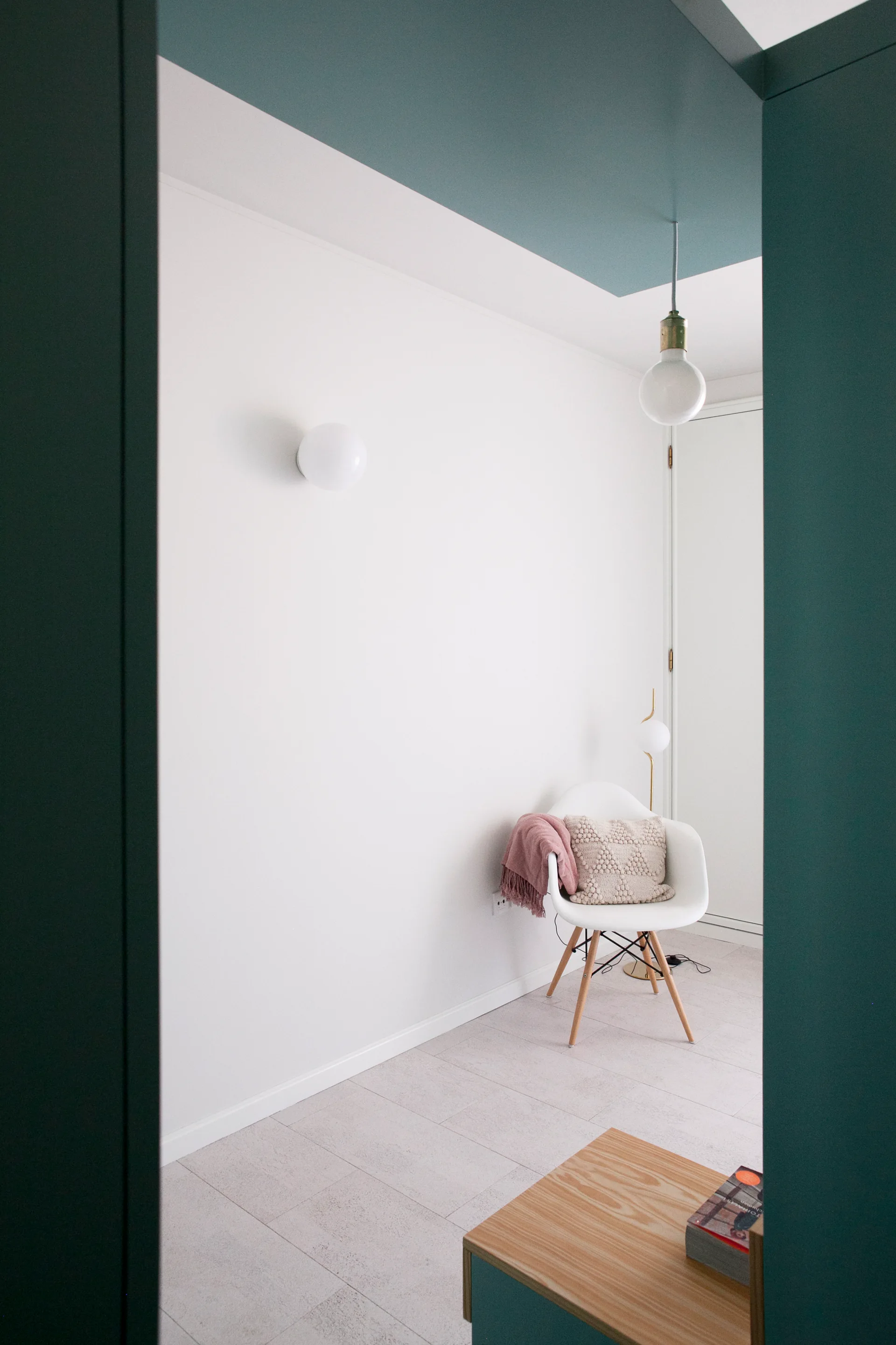 A white armchair with a pink throw sits in a corner beneath a teal painted ceiling and globe light, viewed through a dark teal doorway.
