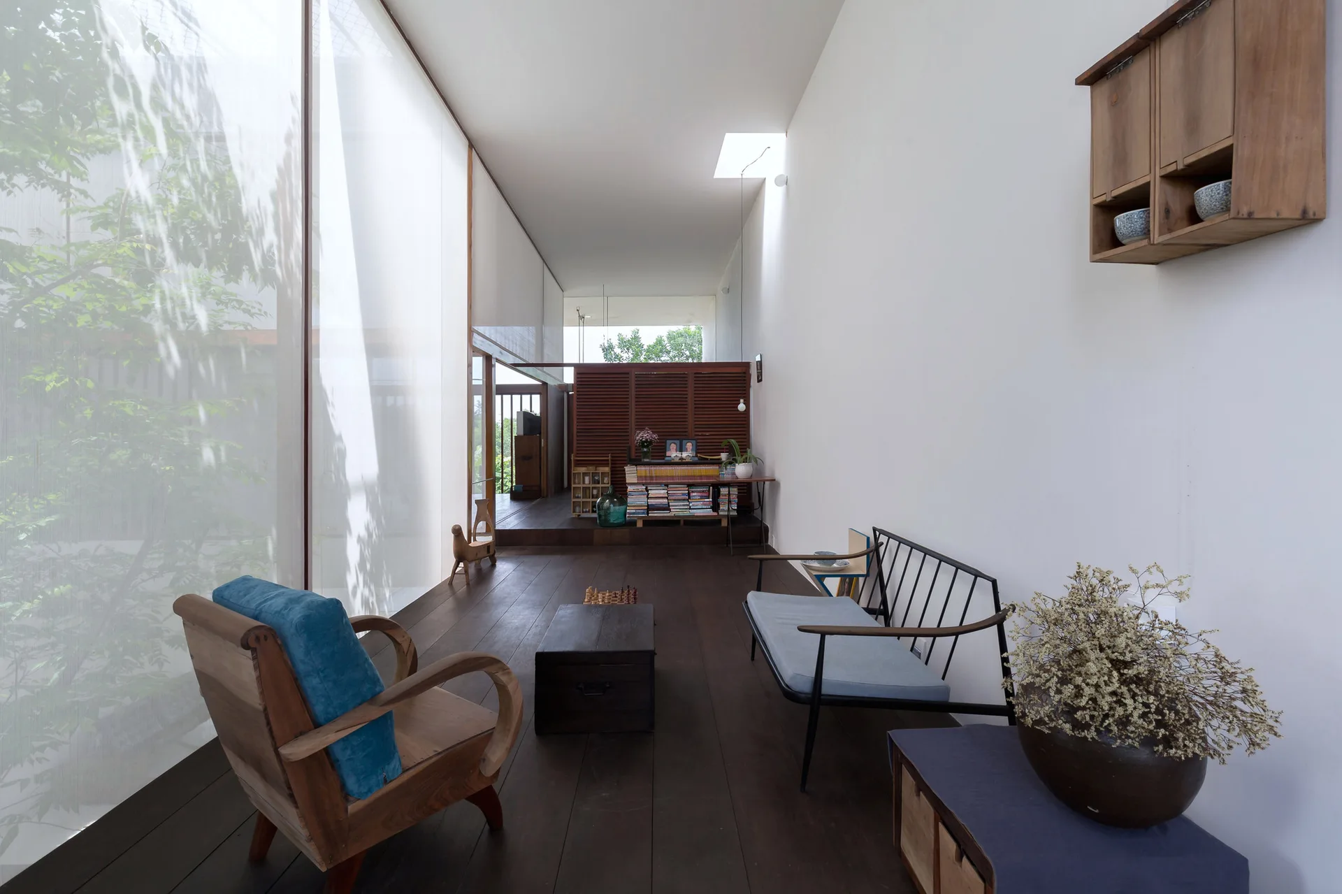 A minimalist living room features dark wood flooring, a blue armchair, a small metal sofa, and a large translucent glass window wall.