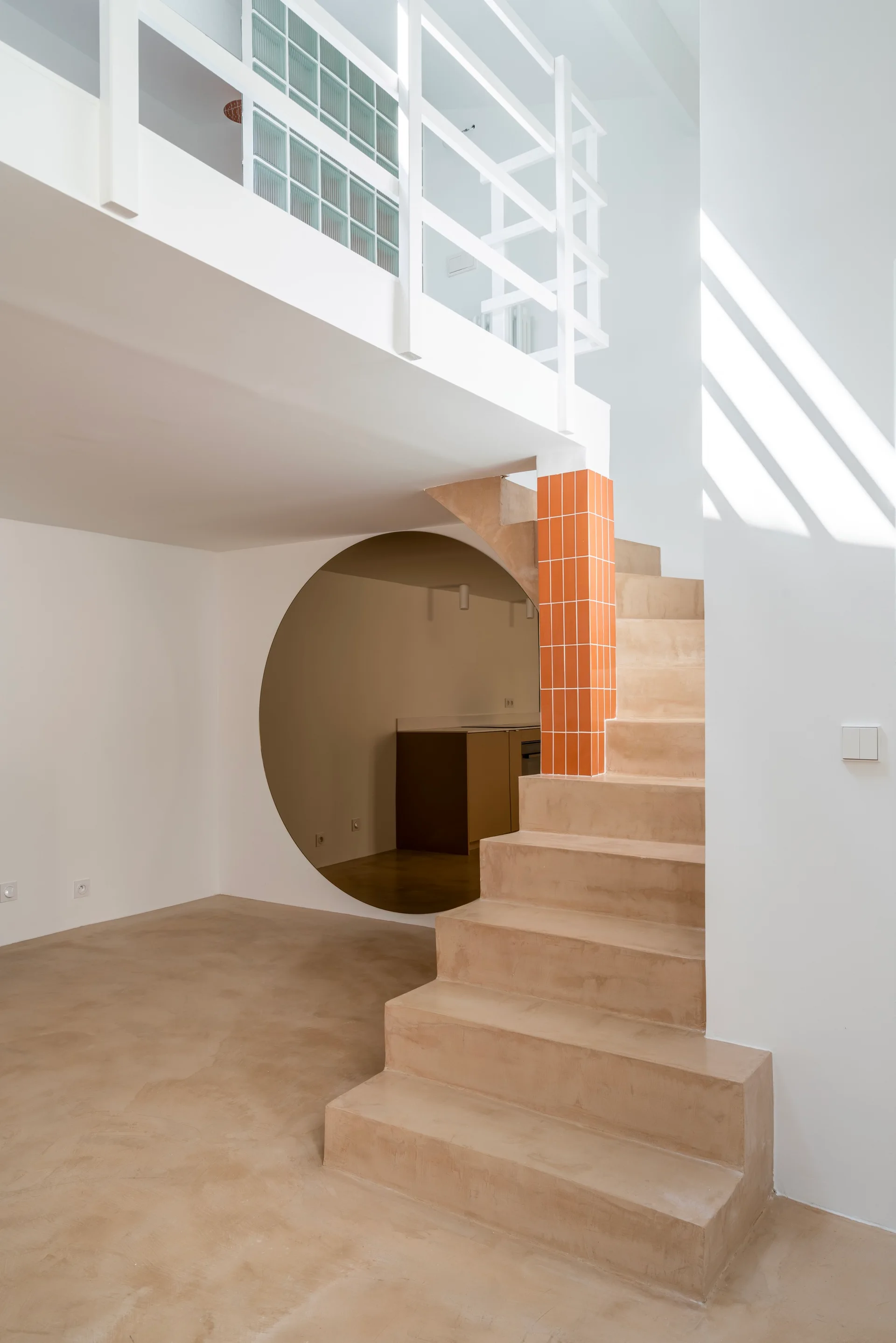 A minimalist interior featuring a sand-colored concrete staircase next to an orange tiled column and a large circular wall cutout.