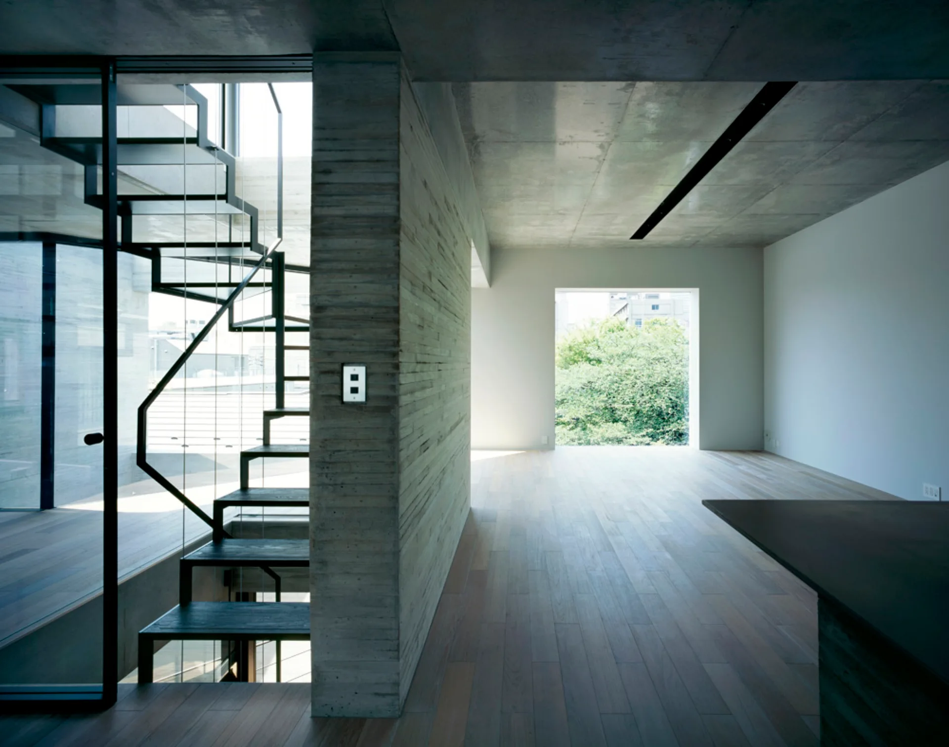A minimalist interior features a black metal open staircase against a textured concrete wall beside light wood flooring.