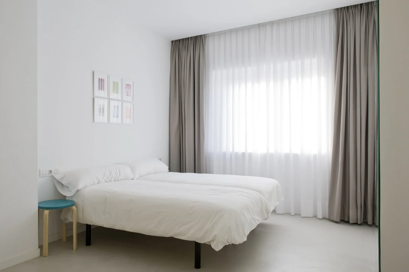 A minimalist white bedroom features a bed with white linens, a grid of small framed art pieces, and grey ceiling-height curtains framing a window.