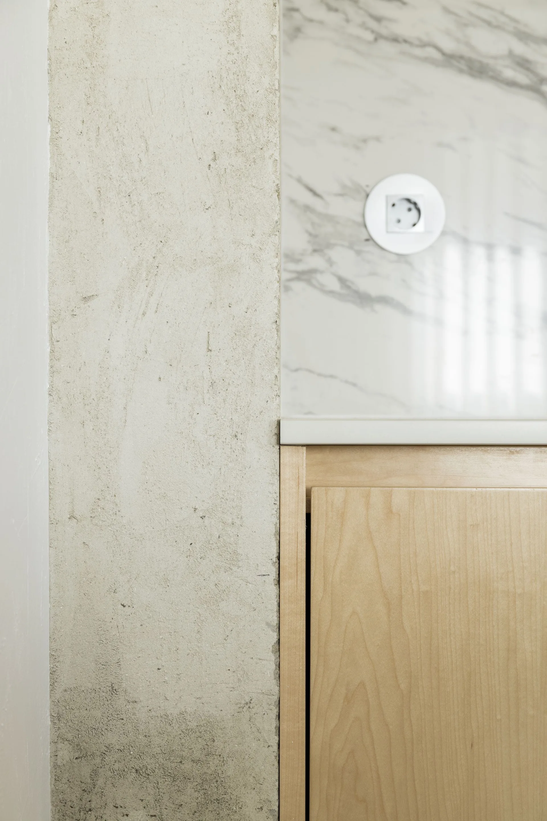 A close-up of a kitchen corner showing a light wood cabinet, a white marble backsplash with a round outlet, and a rough textured concrete wall.