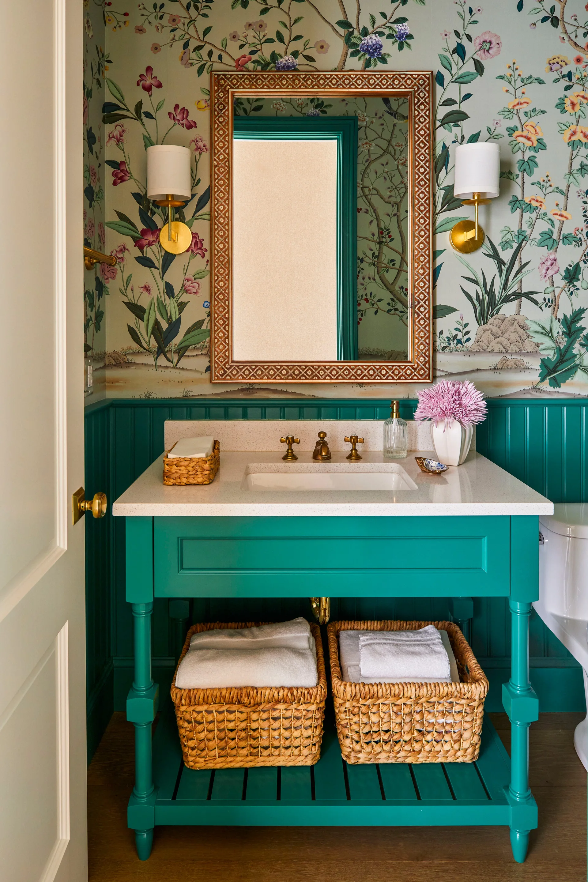 A powder room features a bright teal vanity and beadboard wainscoting below botanical wallpaper, with woven baskets on the lower shelf.