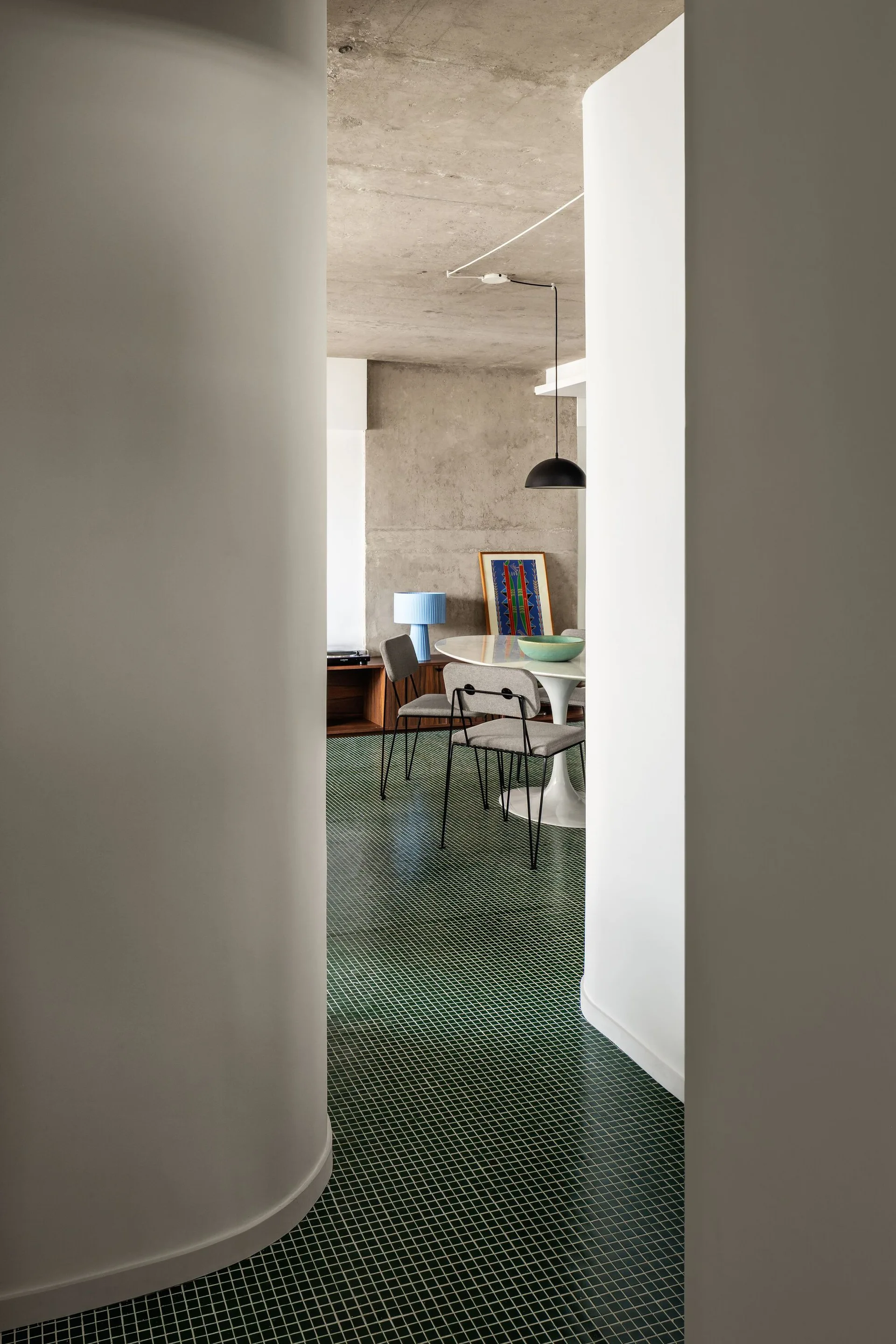 A dining room with green grid-tiled floors, a white pedestal table, and an exposed concrete ceiling, viewed through a curved white doorway.