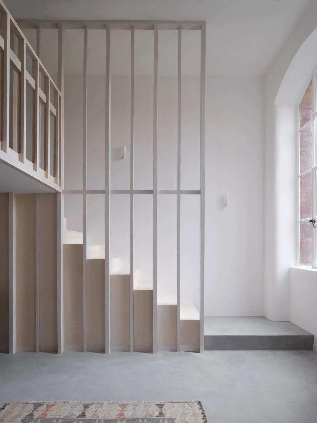 A minimalist light wood staircase is enclosed by a floor-to-ceiling vertical slatted wooden screen on a gray concrete floor next to white walls.