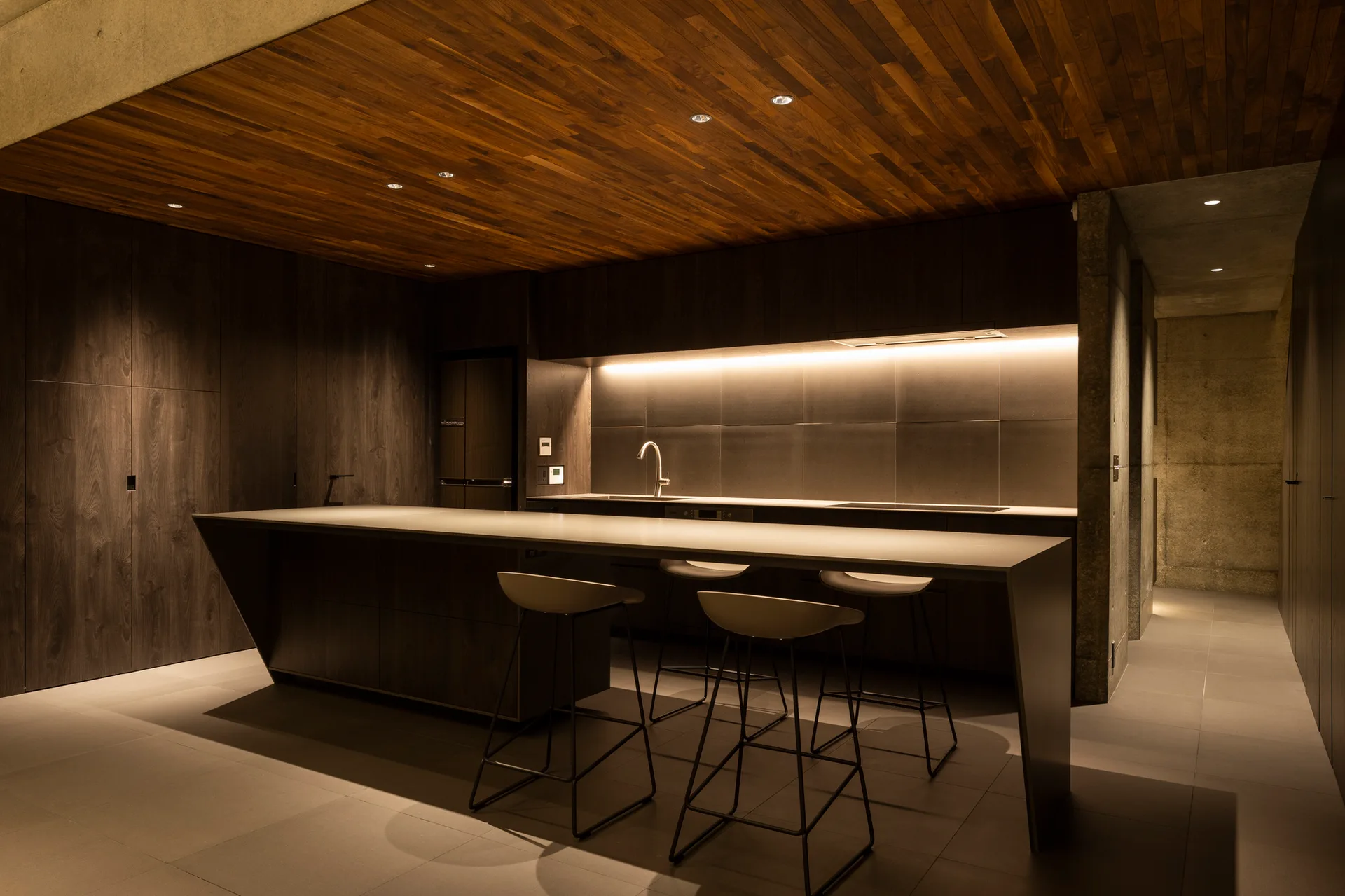 A minimalist kitchen features a large dark island with three bar stools beneath a wood-planked ceiling and warm under-cabinet lighting.