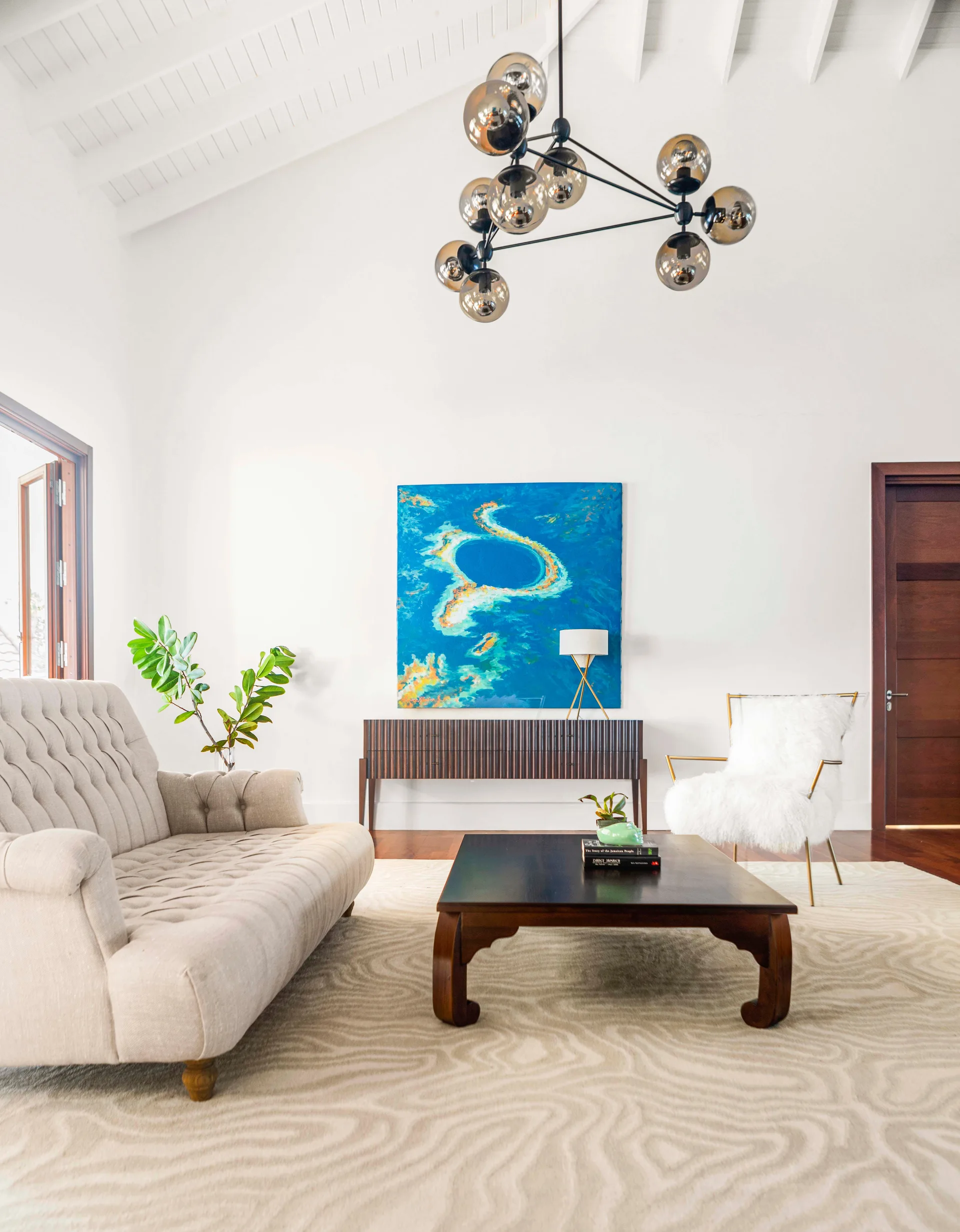 A living room with a white vaulted ceiling, a tufted sofa, a dark wood coffee table, and a large blue abstract painting over a ribbed sideboard.